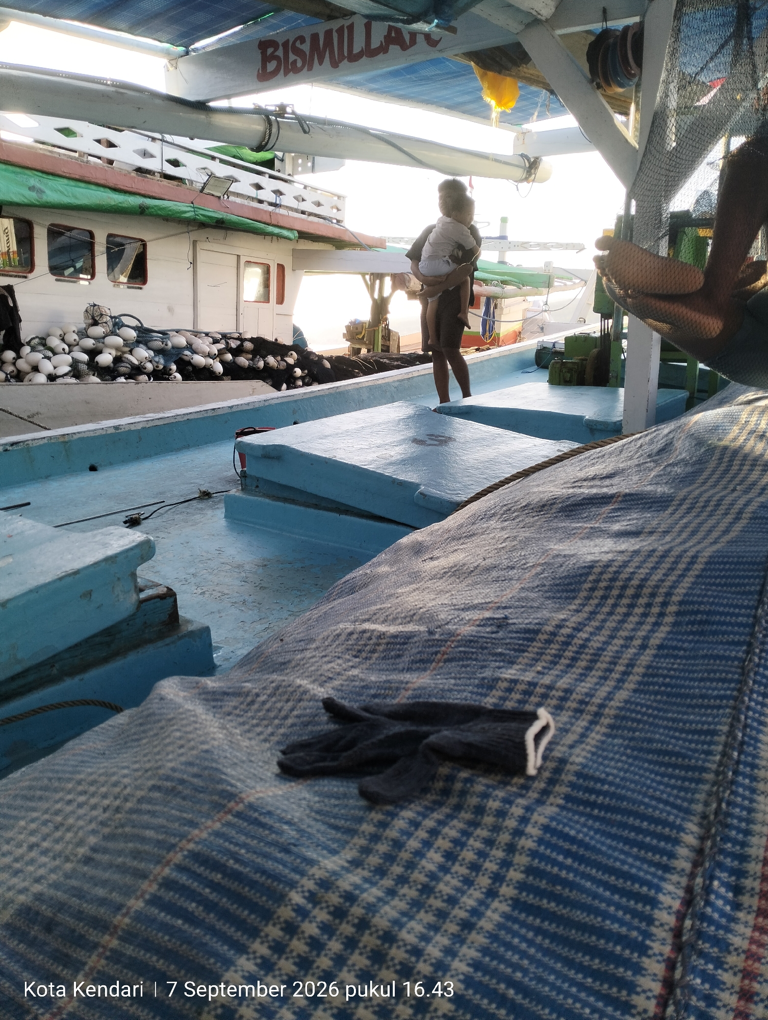 The image captures a view from a boat, likely in the waters of Kendari, Indonesia, as indicated by the timestamp "Kota Kendari | 7 September 2026 pukul 16.43". In the foreground, a blue-and-white patterned blanket or tarp covers a portion of the boat's deck, with a pair of dark gloves resting on top. Behind this, the deck is painted a light blue. Further back, a person is seen holding a young child. The child is wearing a white shirt and a light-colored diaper, while the person is wearing a dark shirt and shorts. Beyond them, a white boat with green accents is docked. This boat has several windows and a stack of white buoys and fishing nets. Overhead, a blue canopy provides shade, and a sign reading "BISMILLA" is visible. The overall scene suggests a moment on a fishing boat at what appears to be late afternoon, given the lighting.