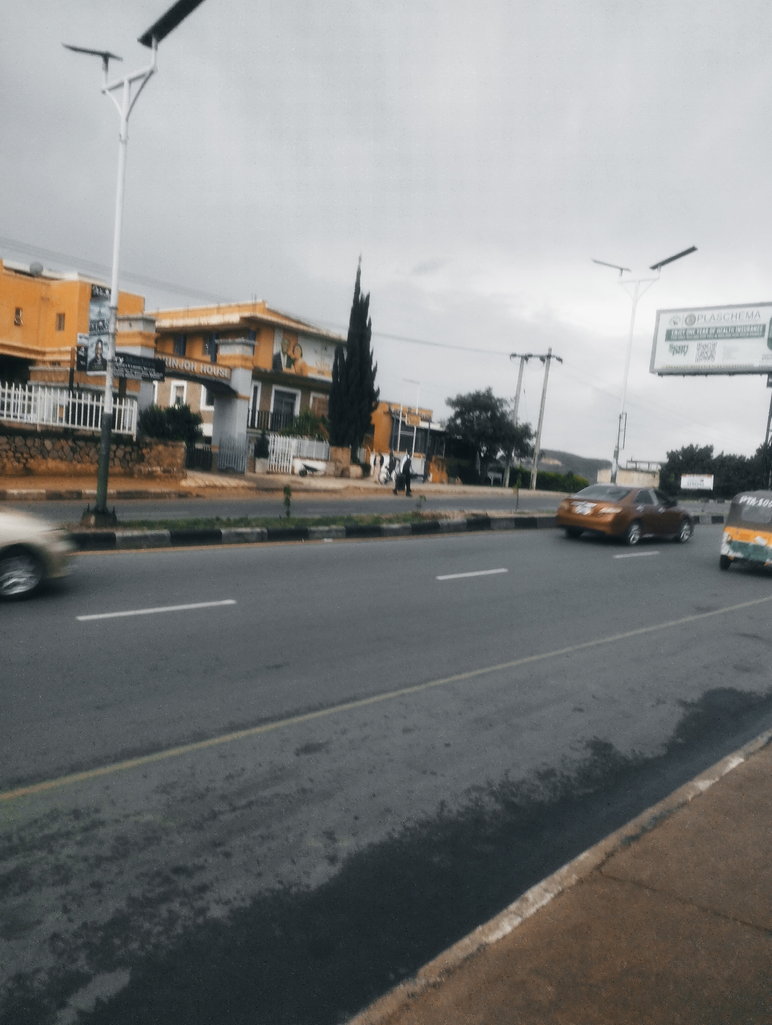 In the city of Jos, Nigeria, on a cloudy day, a view of a street scene unfolds. The foreground is dominated by a gray asphalt road with visible white lane markings. The left side of the road features a strip of black and white checkered curb bordering a sidewalk. A small tree is planted near the curb. In the middle ground, traffic moves along the road. A blurred silver car is seen on the far left, moving away from the camera. To the right, a brownish-orange sedan is driving away from the camera, and a yellow and green three-wheeled vehicle, possibly a tuk-tuk, is also traveling in the same direction.
On the left side of the street, behind a white metal fence, are several buildings. The most prominent is a two-story building painted in shades of yellow and orange. A sign on this building reads "UNION HOUSE." Above the sign, two framed portraits, one of a man and one of a woman, are visible. Another building, partially visible on the far left, is also yellow. A tall, slender dark green conifer tree stands between the buildings. A street light with a solar panel is positioned on the left side of the road.
On the right side of the road, a large billboard is erected. The top of the billboard shows the text "PLASCHEM A." Below this, smaller text reads "ENJOY ONE YEAR OF HEALTH INSURANCE." There are also some QR codes and other smaller text on the billboard. Further down the road, more trees and greenery are visible, and in the distance, hills can be seen under the overcast sky. The overall impression is a typical urban street scene in Jos.