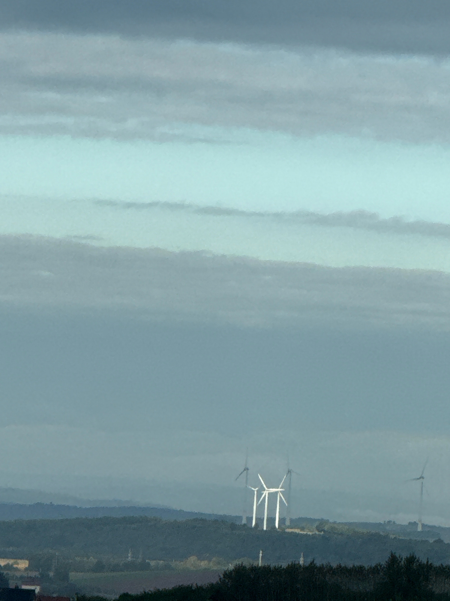 The image depicts a hazy, atmospheric landscape with multiple wind turbines visible in the distance. The foreground features dark, silhouetted trees and some scattered buildings. In the middle ground, rolling hills covered in dense, dark green foliage are visible, with a few structures, possibly a church or tower, peeking through the trees. The background is dominated by a vast sky filled with layers of soft, muted clouds in shades of grey and light blue, creating a diffused light effect. The wind turbines, several of which are grouped together, are tall, white structures with visible blades, some appearing to be in motion. The overall scene suggests a rural or semi-rural environment, possibly on the outskirts of Bad Lauterberg im Harz, Germany, given the characteristic hilly terrain and the presence of wind farms. The visibility is reduced due to the atmospheric haze, lending a somewhat muted and serene quality to the image. No people are discernible in the image.