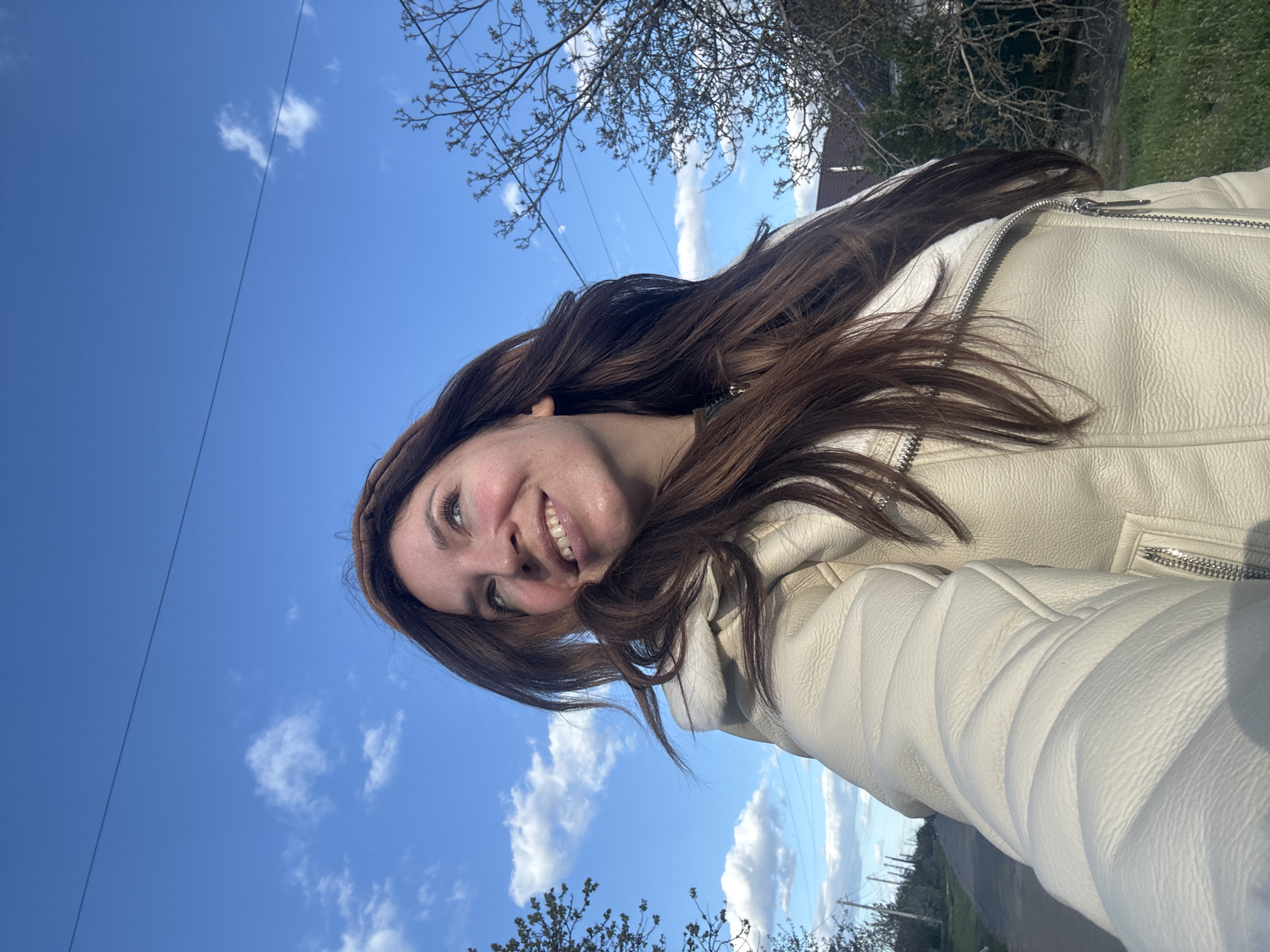 The image captures a young woman smiling brightly, posing for a selfie outdoors. She is positioned in the lower part of the frame, looking upwards towards the camera, with her head tilted. Her long, wavy dark brown hair frames her face and shoulders. She is wearing a light-colored, possibly cream or off-white, leather-style jacket featuring prominent metallic zippers on the front and sleeves, some of which show a quilted pattern.
The background is dominated by a clear, vibrant blue sky dotted with fluffy white clouds, indicating a sunny day. Bare tree branches, characteristic of late winter or early spring, are visible in the upper and lower sections of the frame. A thin, dark utility wire or power line crosses the sky. In the distance, glimpses of green landscape and possibly a building or structure can be seen. The woman's broad smile conveys a sense of happiness and contentment. No visible text is present in the image. The setting is Kryvyi Rih, Ukraine.