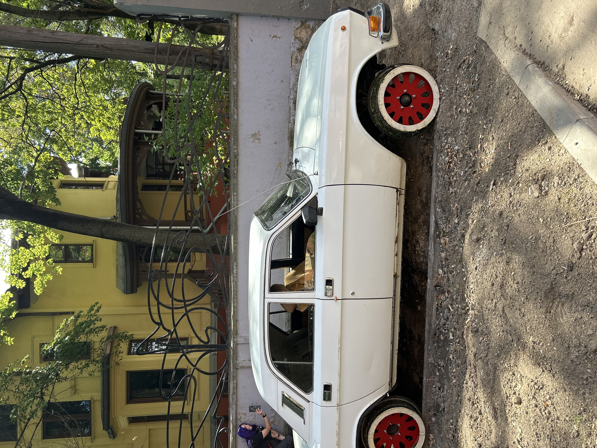 A white vintage sedan with red rims is parked on a dirt road next to a yellow building. A person with purple hair is crouching near the car, taking a picture with their phone. Trees and foliage surround the scene, and a decorative wrought iron fence runs along the property line of the building. The car appears to be in a state of disrepair, with rust visible on the lower body panels.