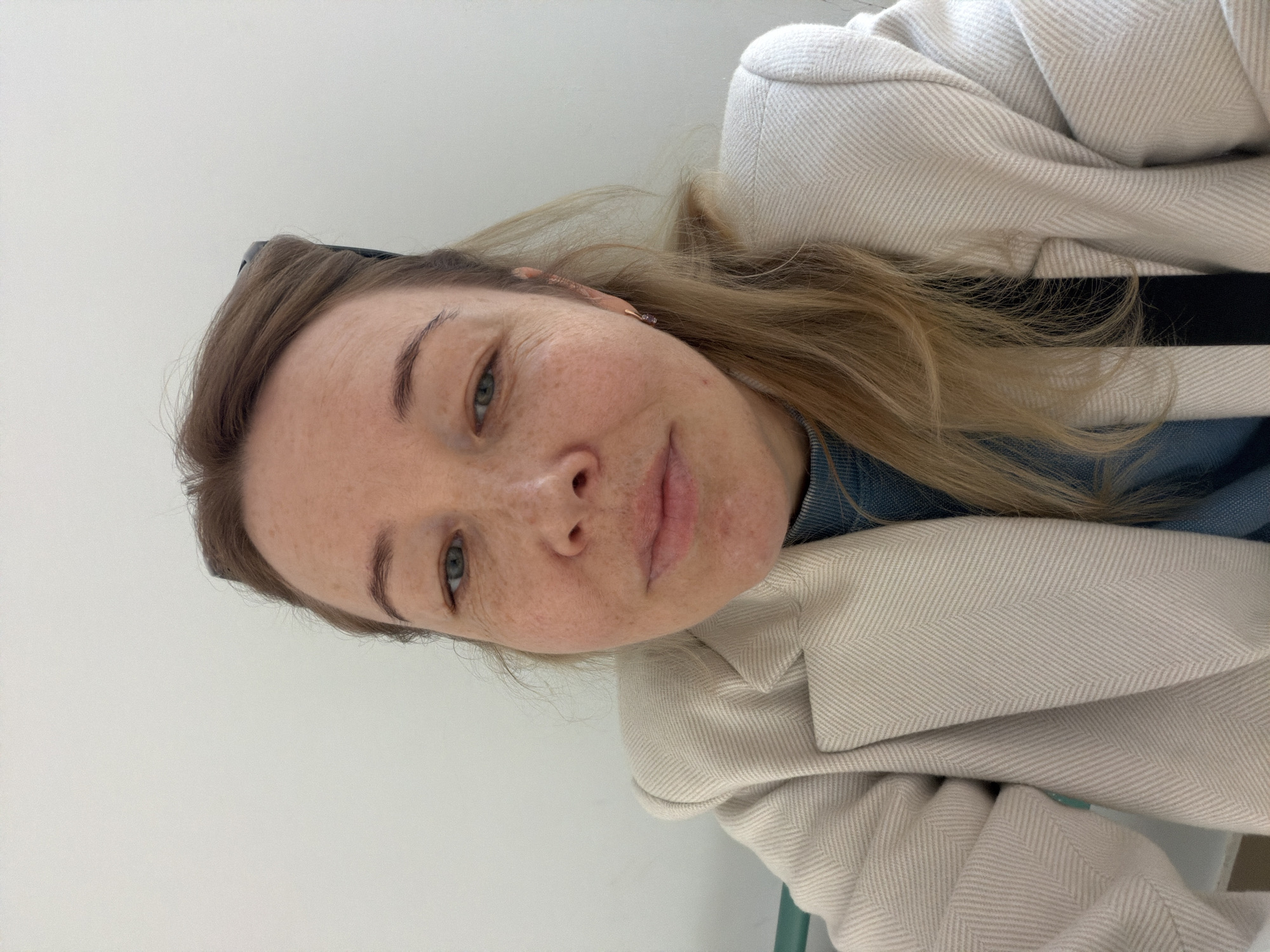 A selfie shows a woman with fair skin, freckles, and light brown hair. She is wearing a blue top underneath a cream-colored jacket with a herringbone pattern. The background is a plain, light-colored wall. There is no information about the location being in the city of Pervomaysky, Russia, in the image.