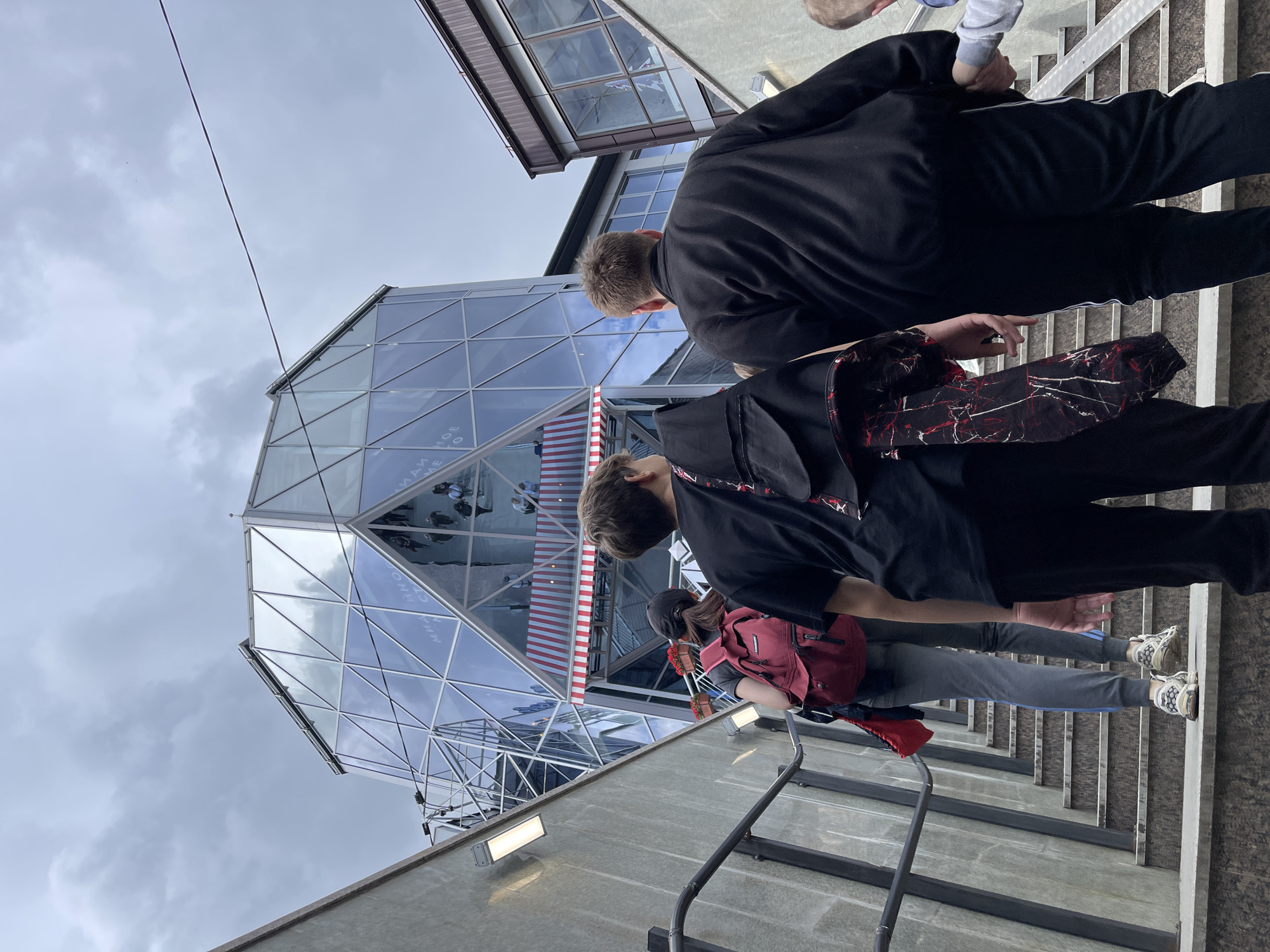 A group of people are descending a staircase next to a unique glass-covered building with a striped awning. The building's exterior is made of triangular glass panels, resembling a geometric design. The setting is Kislovodsk, Russia, based on a visible inscription on the building. People walking on the staircase include individuals wearing casual clothing, with at least one person carrying a red backpack. The weather appears overcast. The stairs have metal railings.