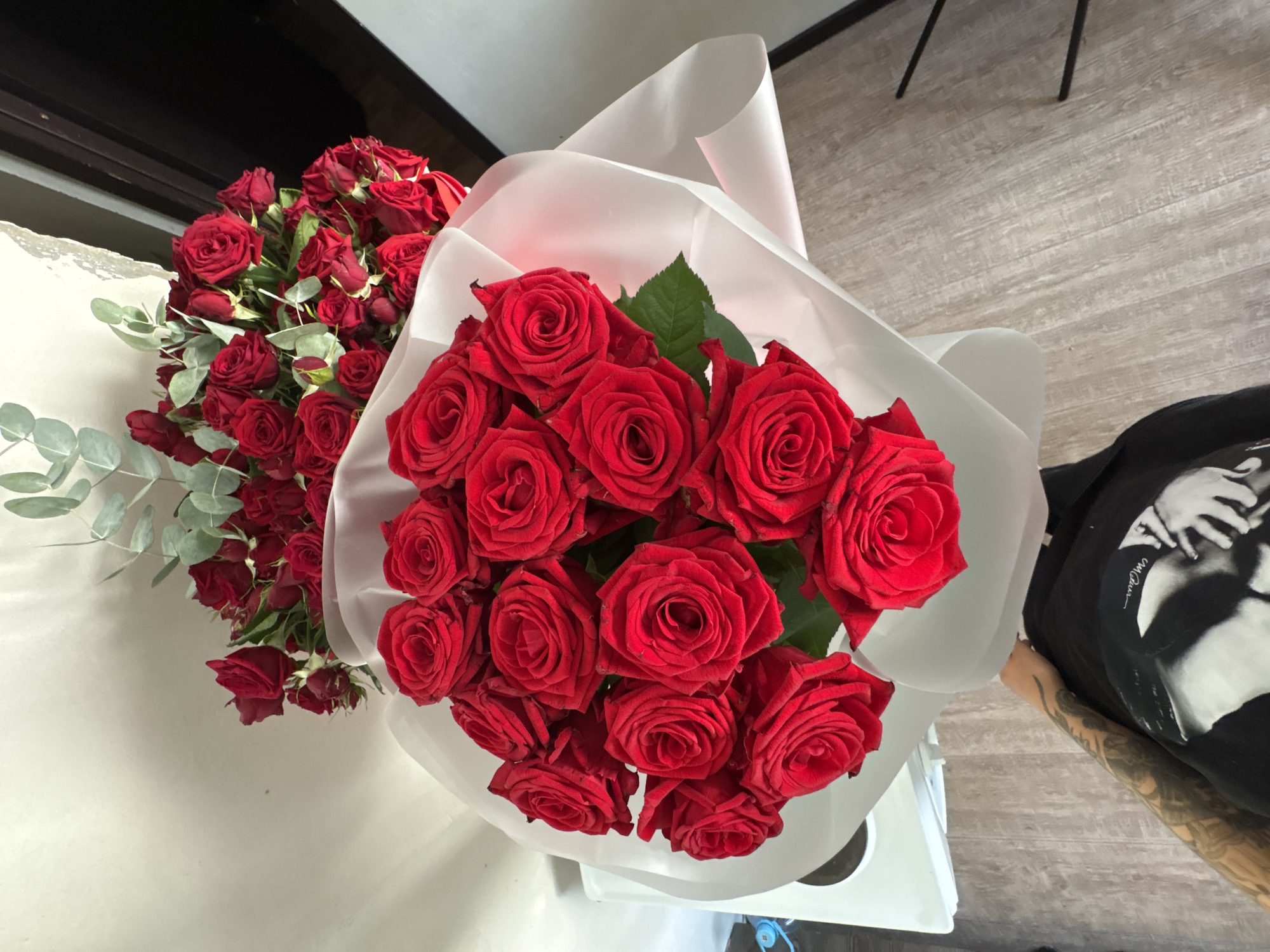 A bouquet of red roses wrapped in white paper is held by a person with tattoos on their arm and chest, wearing a black t-shirt. The bouquet is in the foreground, with another bouquet of red roses and eucalyptus in the background. The scene appears to be indoors, with a light-colored wall and a wooden floor visible. The lighting suggests it could be daytime, with natural light casting shadows. There is no indication of the specific location within Rostov-on-Don, Russia.