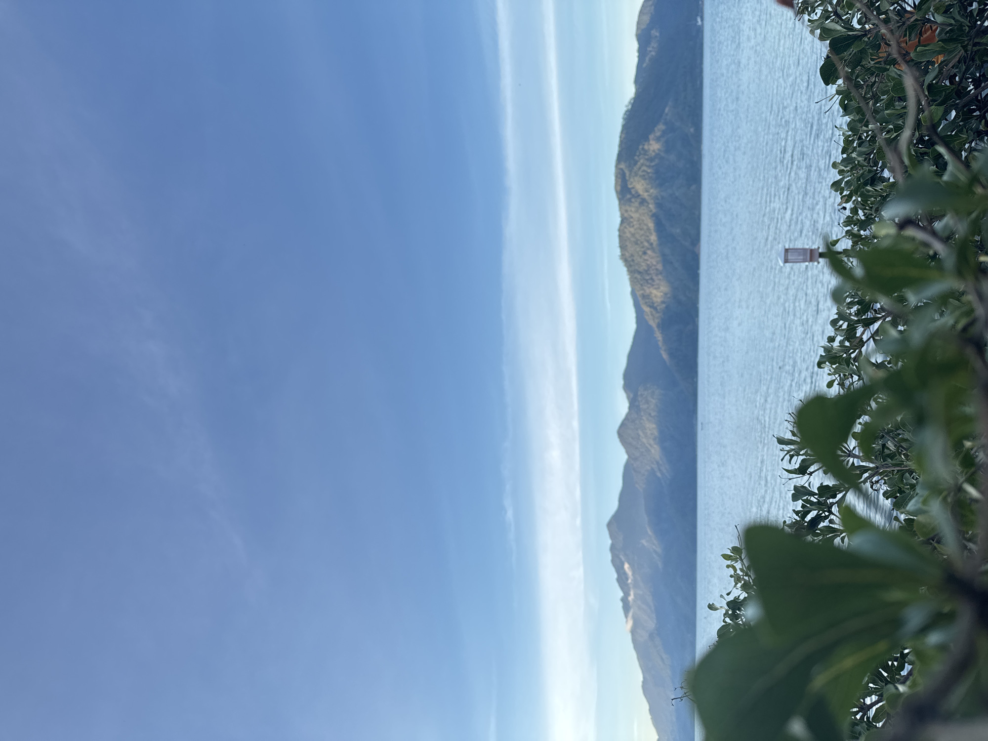 An outdoor, low-angle, vertical shot captures a tranquil lake scene in the city of Kalalengkio, Indonesia, under a bright, clear blue sky with wispy white clouds. In the foreground, lush green foliage partially obscures the view, with some leaves in sharp focus while others are blurred. Beyond the foliage, a calm, light blue lake stretches across the middle ground, its surface rippled by a gentle breeze. In the distance, a range of majestic, green mountains rises, their peaks bathed in the soft light of what appears to be early morning or late afternoon. A small, white lantern with a brown base is visible in the mid-ground, positioned near the water's edge. The overall atmosphere is serene and picturesque.