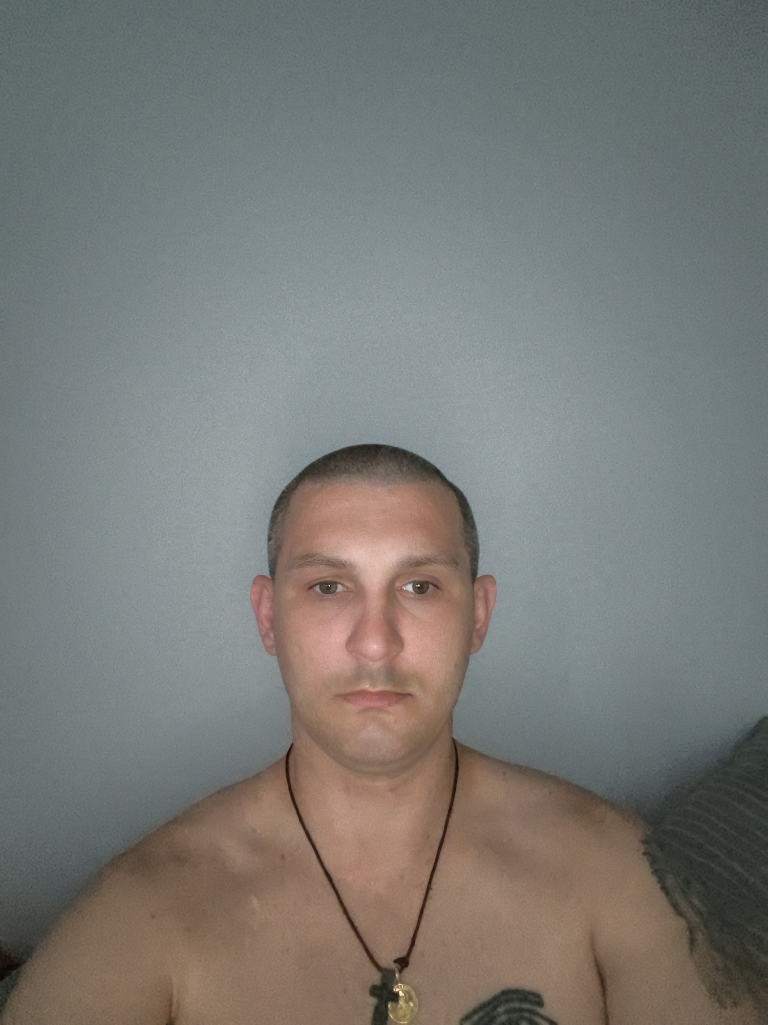 The image is a close-up, head-and-shoulders portrait of a shirtless man with a buzz cut. He has light brown eyes, a neutral expression, and is wearing a dark necklace with a pendant. There are tattoos visible on his chest and upper left arm. The background is a plain, light grey wall. The man is positioned in the center of the frame. The overall lighting is dim, with the light source appearing to be from the front and slightly above the subject. There is a subtle texture to the wall, suggesting it is painted or a similar material. No specific location in Wolgast, Germany can be determined from the image.