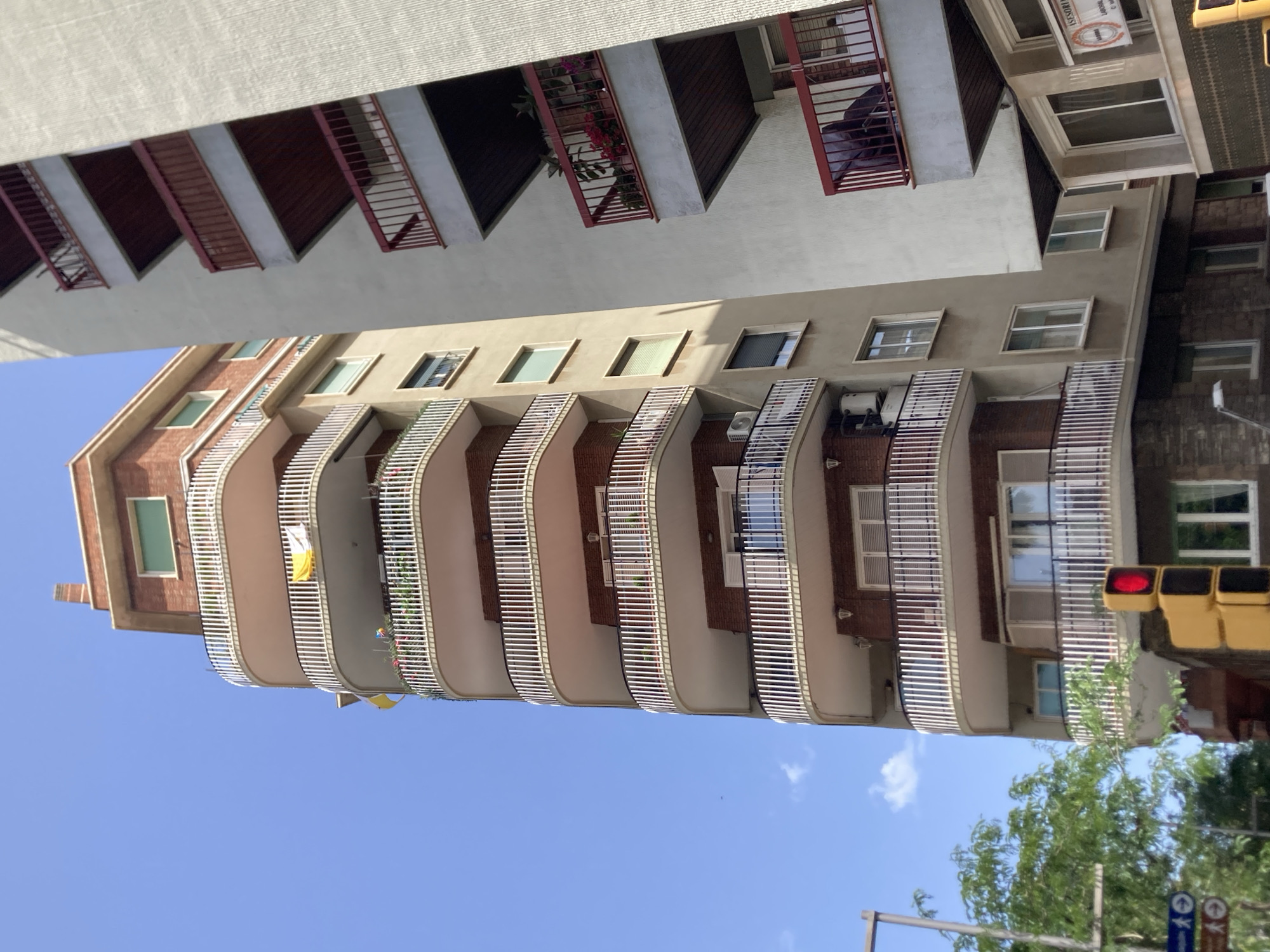 The image shows an outdoor, daytime, low-angle view of two apartment buildings in Barcelona, Spain. The building on the left is a light brown, curved building with multiple balconies featuring white railings and pink overhangs. The building on the right is a cream-colored building with red-railed balconies. The sky is a clear blue with a few wispy clouds. In the lower left corner, a traffic light with a red signal is visible, along with some green trees and street signs. The scene appears to be a typical urban streetscape.