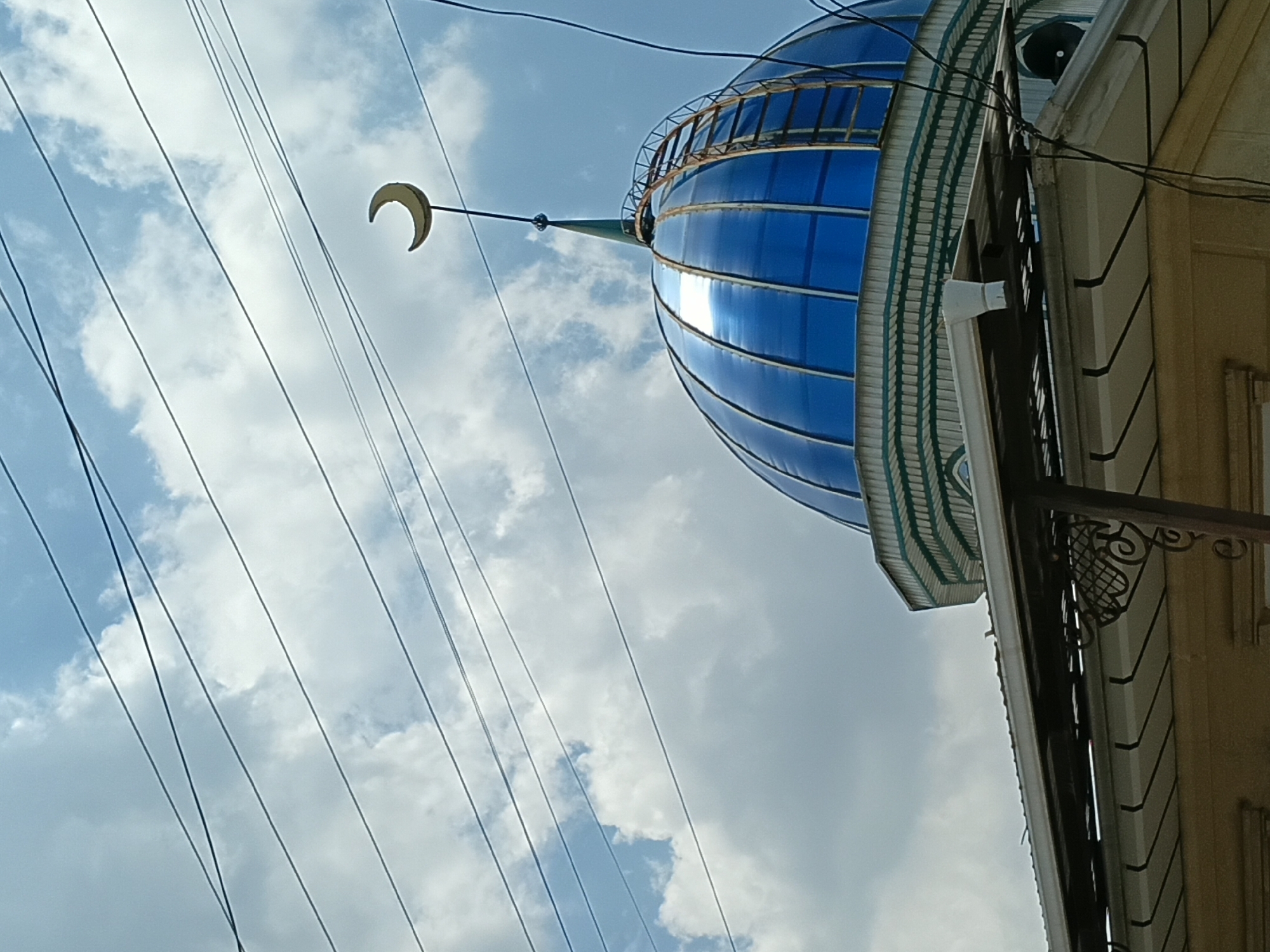 The image shows the blue dome of a mosque, topped with a crescent moon symbol, against a cloudy sky crisscrossed by power lines. A rusty ladder is attached to the side of the dome. The lower part of the image reveals the eaves of a building, with decorative metalwork and a portion of the building's facade visible. The scene is likely outdoors, possibly in the city of Osh, Kyrgyzstan, given the presence of a mosque. No people are visible in the frame.