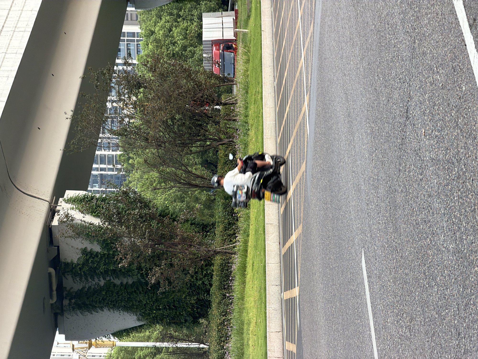 A person wearing a white shirt and black shorts is riding a black scooter on a road in the city of Jiangjin, China. The scooter has a storage box on the back. The road has yellow and white markings. In the background, there is a grassy area with trees and buildings. A large concrete structure with ivy growing on it is visible, as is a red truck carrying white panels. The scene appears to be under an elevated road or bridge.