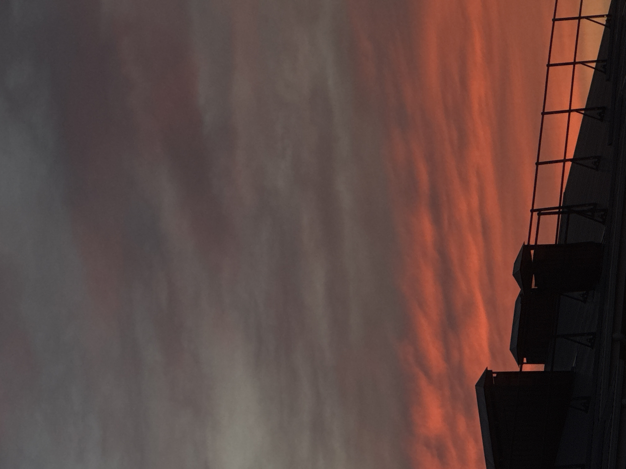 The image captures a section of a building's rooftop in Mikhaylovsk, Russia, against a dramatic sunset sky. The sky is filled with wispy clouds, predominantly in shades of orange and red near the horizon, transitioning to darker grays and purples higher up. Silhouetted against this vibrant sky are three dark structures on the roof, likely chimneys or ventilation shafts, each with a distinct cap. A dark railing runs along the edge of the roof, also silhouetted. The rooftop itself appears to be dark and possibly sloped. There are no people visible in the image. The overall impression is one of a peaceful evening scene in a Russian city.