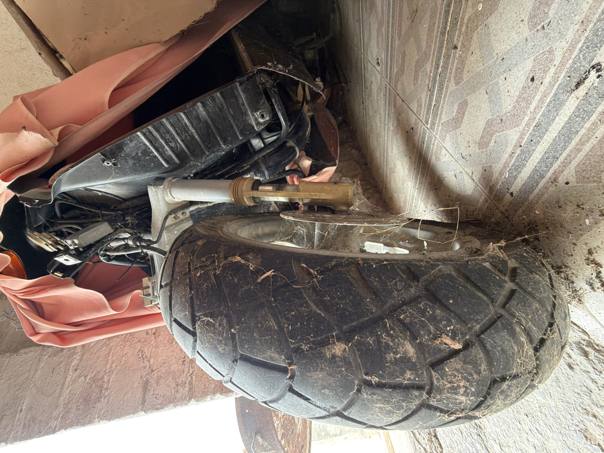 The image depicts a neglected motorcycle in a cluttered, seemingly indoor environment. The front tire is prominently displayed, covered in dirt and spiderwebs, suggesting prolonged disuse. Part of the motorcycle is draped with a faded pink cloth. The background reveals a textured wall with a patterned design, possibly tile or patterned paint, and a concrete wall. The scene suggests the motorcycle is in storage, possibly in a garage or shed, with no visible people or activity.
There is no information to identify this location as in the city of Sant Martí Sarroca, Spain.