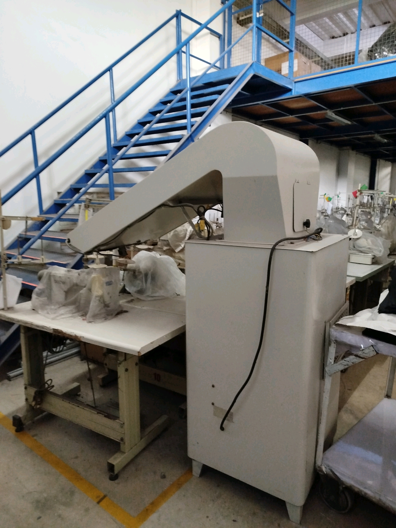 This image depicts an indoor industrial setting, possibly a workshop or factory floor, with elements suggesting manufacturing or production. In the foreground, a large, light-colored industrial machine with a curved top section dominates the right side of the frame. A black electrical cord trails from this machine towards the ground. To the left of this machine, a white workbench is positioned. On the workbench, several industrial sewing machines, covered in protective plastic, are visible. A blue metal staircase with railings rises from the lower left of the frame towards an upper level of the building, which appears to be a mezzanine with metal grating flooring and blue metal railings. Boxes are stacked on the mezzanine. The environment is utilitarian, with concrete flooring marked with yellow lines. Other industrial equipment and machinery, as well as some hanging items that could be textiles or materials, are visible in the background, suggesting ongoing operations. The specific location of Surakarta, Indonesia, is not explicitly identifiable from the visual cues alone, but the presence of industrial equipment and the general layout are consistent with a manufacturing facility. There are no people visible in the image.