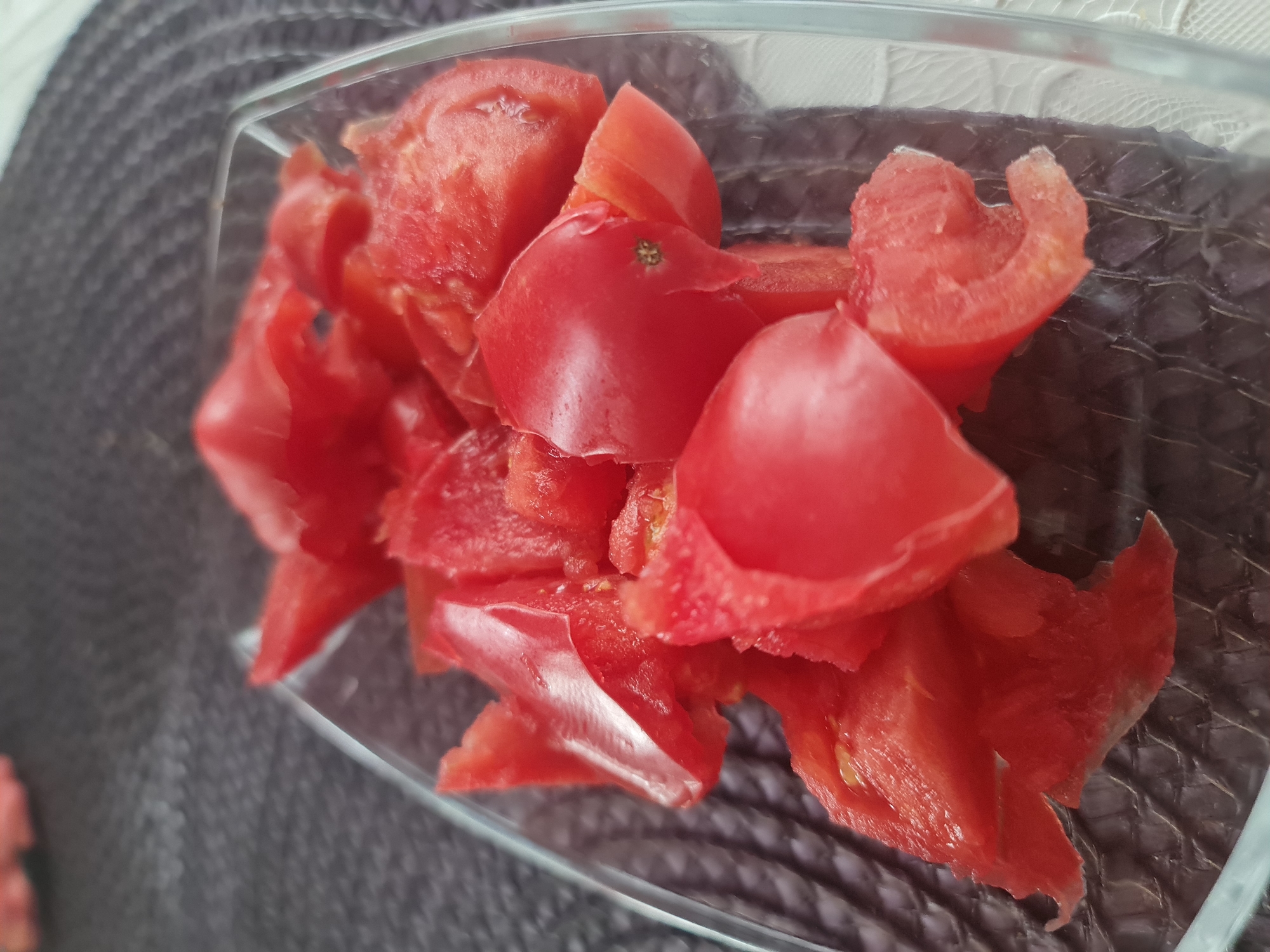 A close-up, high-angle shot shows chopped red tomatoes in a clear rectangular glass dish. The tomatoes are unevenly cut and appear fresh. The dish is placed on a dark, textured placemat, possibly made of woven material, with a hint of white lace or fabric visible on the right edge of the frame. The background is softly blurred, indicating a shallow depth of field. There are no people or animals present in the image. The scene is indoors, likely a domestic setting. The city of Angarsk, Russia, is not depicted in this image.