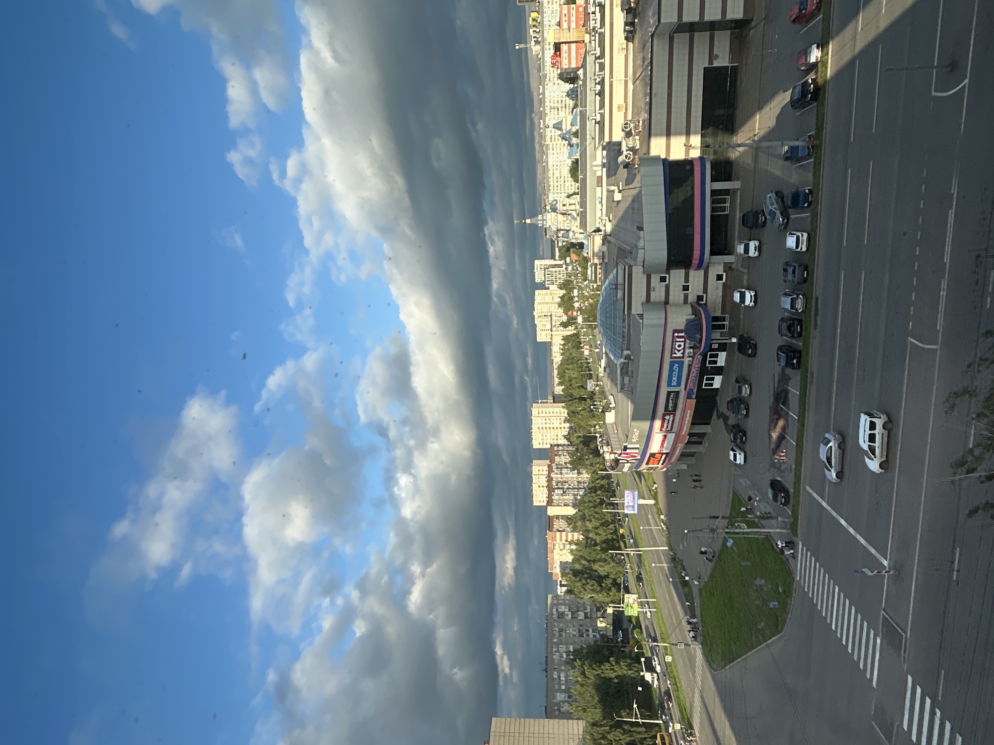 The image shows a view from above of a city intersection and a shopping mall in Izhevsk, Russia. The sky is blue with a mix of white and dark clouds. The shopping mall, identified by signage as "SOKOLOV KART", is a modern building with a domed section and a prominent pink and blue stripe. In front of the mall, there is a parking lot with several cars.
The intersection is well-lit and features a pedestrian crossing. Cars are seen driving and parked in the vicinity. To the left of the intersection, there is a grassy embankment with trees lining a street that leads to residential buildings. These buildings are multi-story, typical of urban housing in Russia. Further in the distance, more residential buildings and a church with blue domes are visible. The overall impression is a typical urban scene on a partly cloudy day.
