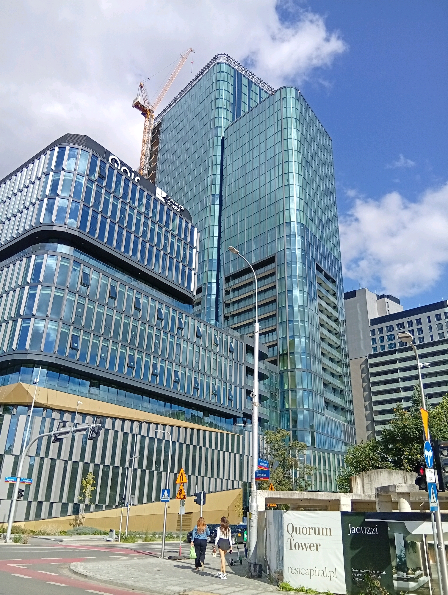 The image depicts a street scene in Wrocław, Poland, specifically at the Quorum complex. In the foreground, two women are walking on a sidewalk with a construction site to their right. The construction site features a large banner advertising "Quorum Tower" and "Jacuzzi" with a website address. The background is dominated by modern glass-fronted buildings, including the Quorum Tower itself, which is a tall skyscraper. A construction crane is visible atop one of the buildings, indicating ongoing development. The sky is blue with scattered clouds, and streetlights and traffic signals are present. The architecture is characteristic of contemporary urban development, with a focus on glass and steel structures.