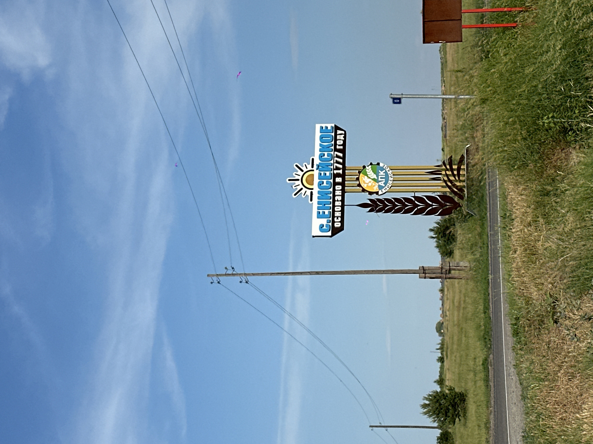 An outdoor, eye-level, medium shot shows a sign for the city of Eniseiskoe, Russia, on a bright, sunny day with a blue sky and wispy clouds. The sign, located in the foreground to the right, is tall and features the words "С.ЕНИСЕЙСКОЕ" (S. ENISEISKOE) in large, blue, capital letters with a stylized sun image above. Below the city name, in smaller text, it reads "ОСНОВАНО В 1777 ГОДУ" (FOUNDED IN 1777). The sign also includes a circular emblem with the letters "АПК" and smaller text that appears to be "ЕНИСЕЙСКОЕ." The sign is mounted on a structure resembling stalks of wheat.
To the left of the sign, a tall wooden utility pole with several wires stretches across the frame against the sky. Further to the left, another similar pole is visible in the distance. A paved road runs across the lower portion of the image, bordered by green grass and vegetation. The background consists of a large, flat, grassy field with scattered trees and some distant structures. Two small pink kites are visible flying in the sky, one to the left and one to the right of the main sign. To the far right of the sign, a brown rectangular sign with red posts is partially visible. The overall environment appears rural.