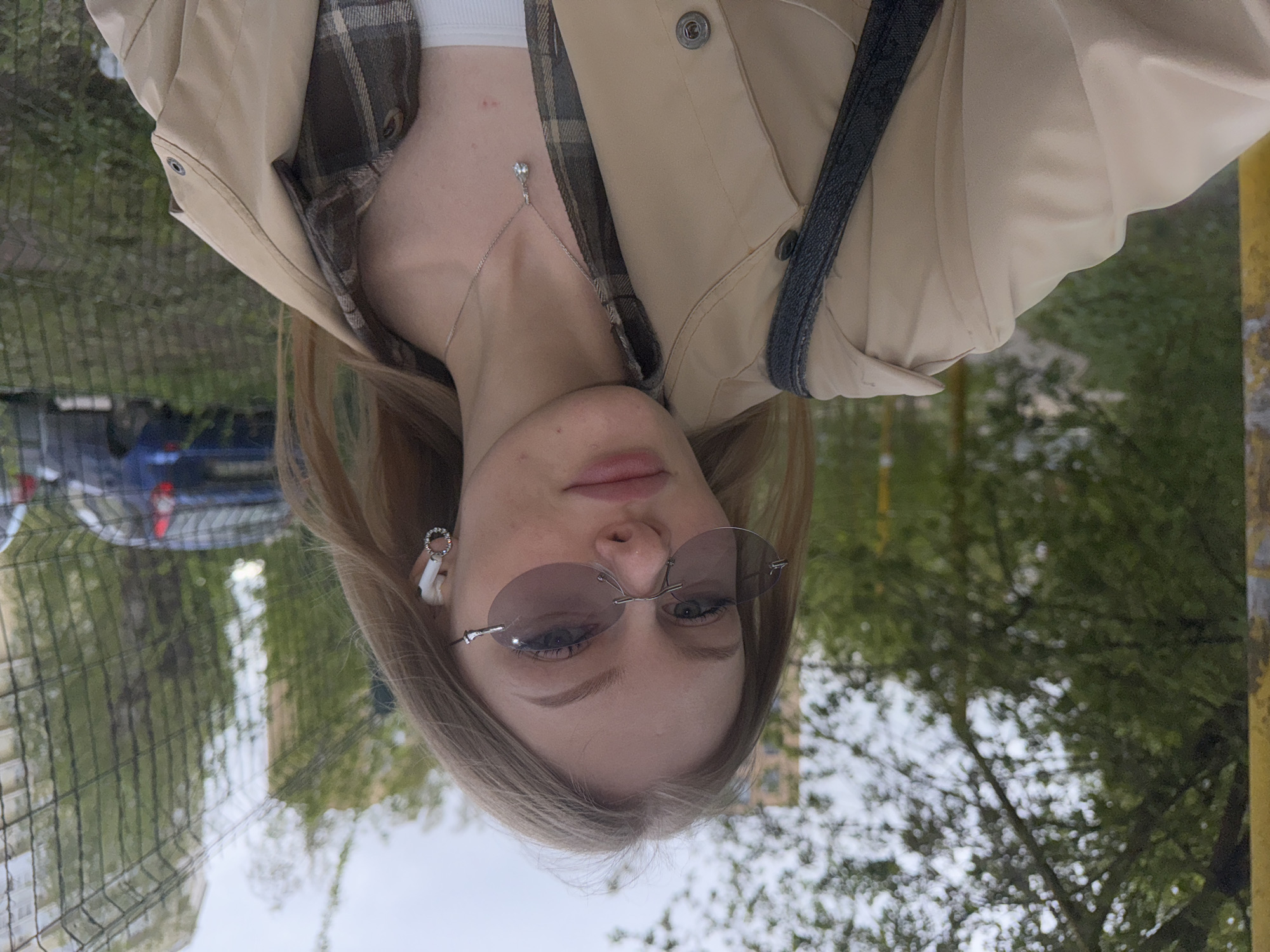 A young, fair-skinned woman with blonde hair is the subject of the photograph, taken from a slightly upward angle. She is wearing rimless sunglasses, a white earpiece in her left ear, and a delicate necklace with a pendant. She is also wearing a combination of clothing: a brown plaid shirt and a beige jacket with a dark gray shoulder strap. The background features a chain-link fence, trees with green foliage, and a glimpse of a blue car. The location appears to be an outdoor urban area. The photo's orientation is slightly tilted. The precise location in Perm, Russia is not identifiable from the image.