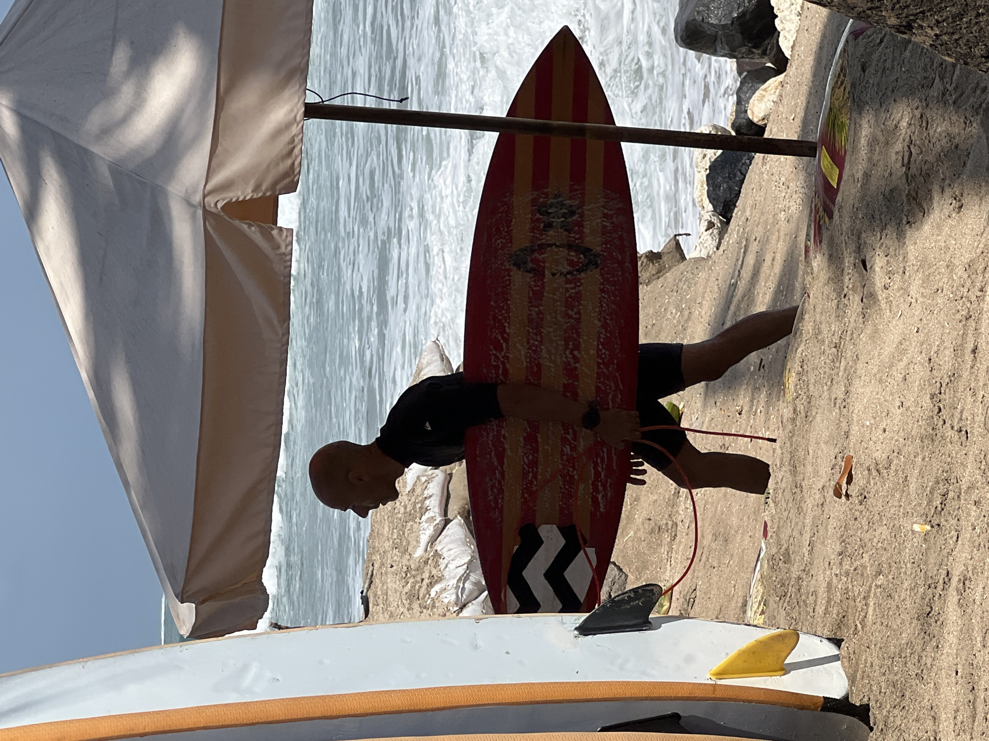 A man in a black wetsuit carries a red and yellow striped surfboard down a sandy beach in Kuta, Indonesia. He is walking from left to right, with his profile visible as he looks out at the ocean. The surfboard has a black and white zig-zag pattern on the tail. In the foreground, another surfboard stands upright. A large, light-colored umbrella provides shade over the beach. The ocean is visible in the background with moderate waves. The scene is set on a sunny day.