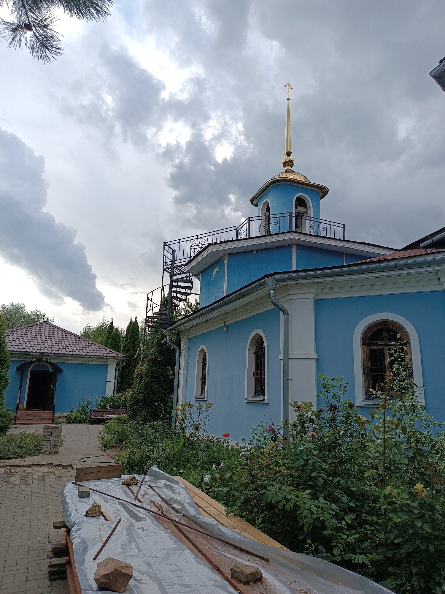 The image shows the Church of the Ascension of the Lord in Bykovo, Russia, under a cloudy sky. The church is a striking blue with white trim and features a golden dome topped with a cross. A spiral staircase is attached to the exterior of the main building. Adjacent to the church, a smaller building with a brown roof is visible, with an open doorway. The foreground displays construction materials covered with a gray tarp, including wooden planks and metal rods, along with scattered rocks. Lush green bushes and flowers are planted around the buildings. The ground is paved with bricks.