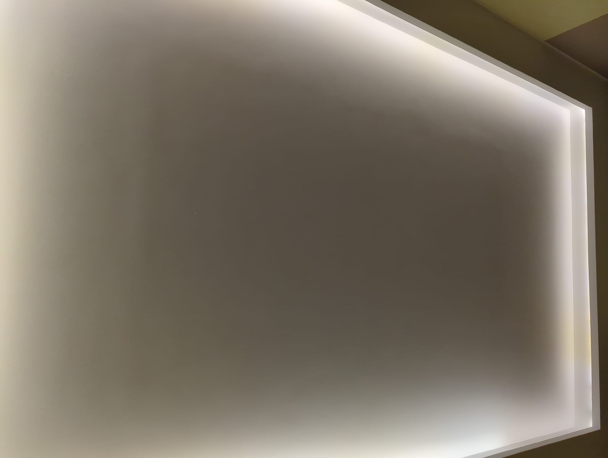 The image shows an interior architectural detail, likely a section of a wall or ceiling. The surface is a matte, pale color, possibly white or light gray. A recessed light fixture or a similar design element frames the surface, casting a bright, diffused glow around the edges. The light is brightest along the perimeter, gradually fading towards the center of the surface. There is no immediate context to indicate the location as Minsk, Belarus, or to determine any specific actions, interactions, people, or objects.