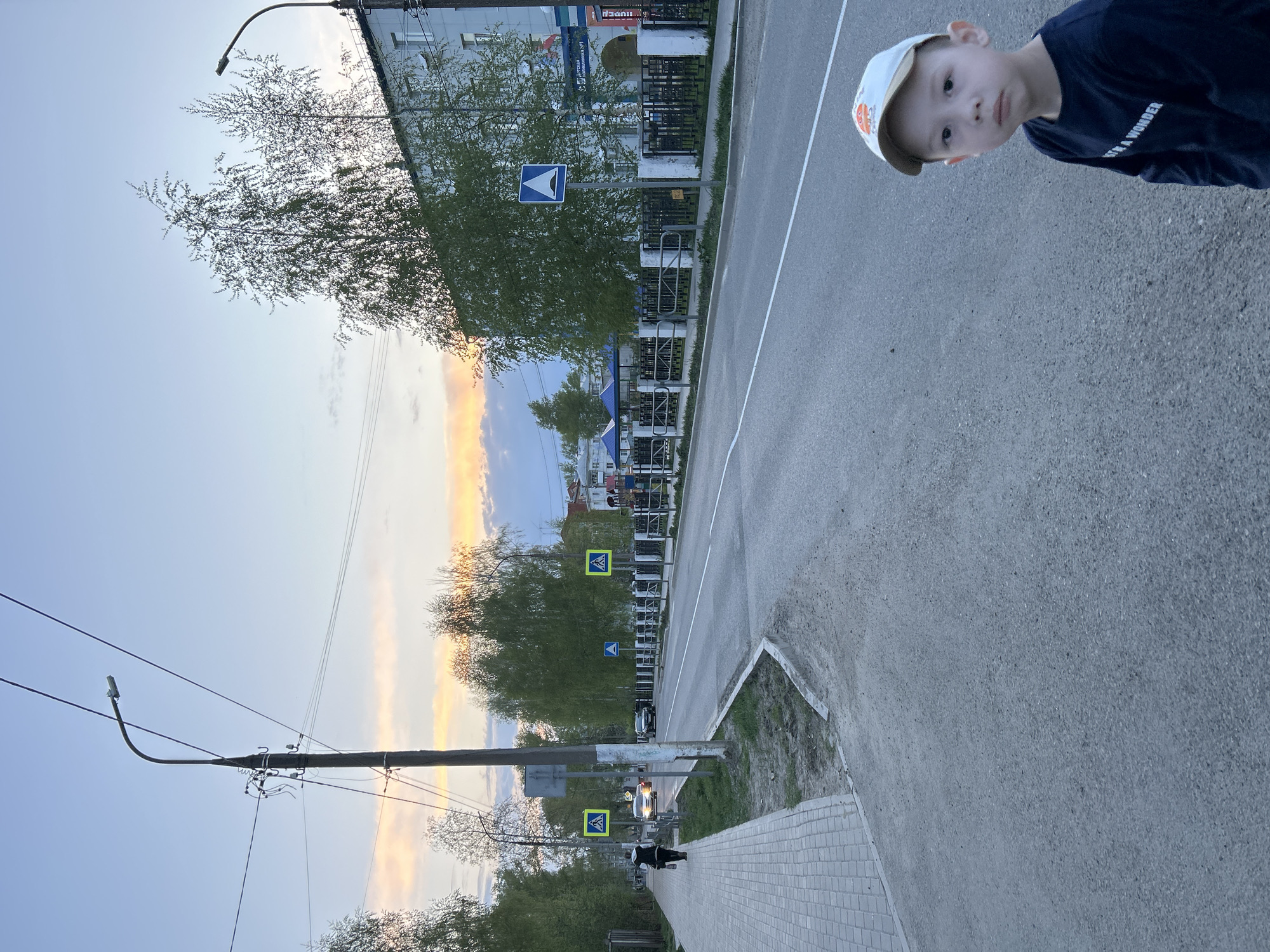 A young boy is standing on a paved sidewalk in Chaikovsky, Russia. He is wearing a blue shirt and a light-colored baseball cap. The sidewalk is next to a road with a white line down the middle. There are pedestrian crossing signs on both sides of the road. There are trees and buildings in the background. The sky is a mix of blue and orange, suggesting it is either sunrise or sunset. Power lines and street lights are also visible.
