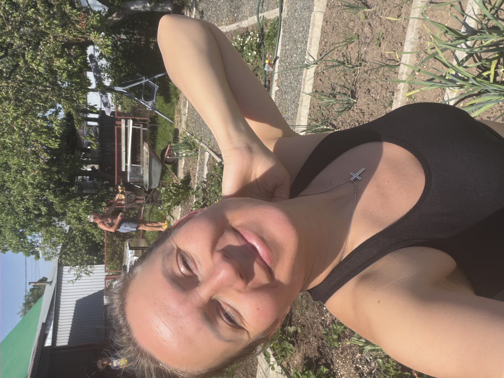 A woman takes a selfie outdoors in Chaikovsky, Russia. She has her arm raised behind her head, is wearing a black tank top and a silver necklace with a cross pendant. Behind her is a garden with what appears to be growing onions and a man standing near a wheelbarrow. In the background is a building with a green roof and another building further away.