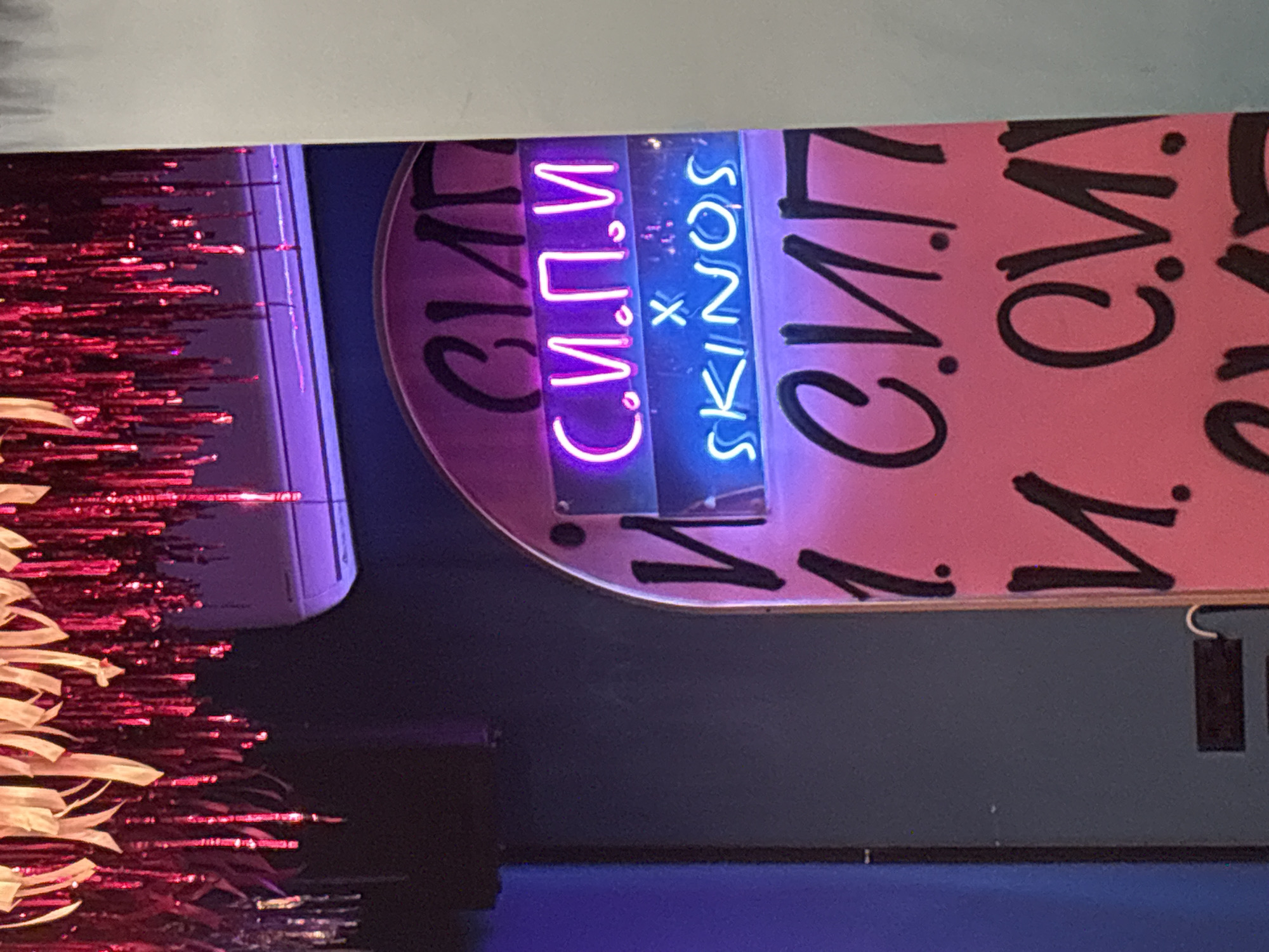 The image shows a portion of a vertical sign for "C.V. SKIVOS," likely a business or organization. "C.V." is represented in Cyrillic lettering in the upper part of the sign using a purple neon light. Below that, “SKINOS” is written in blue neon lighting. The sign is made of a pink background. The pink background has Cyrillic letters written in black. The sign is mounted on a dark gray wall. To the left of the sign are strands of decorative tinsel. The image was likely taken inside. The location in the city of Plovdiv, Bulgaria, is not known.