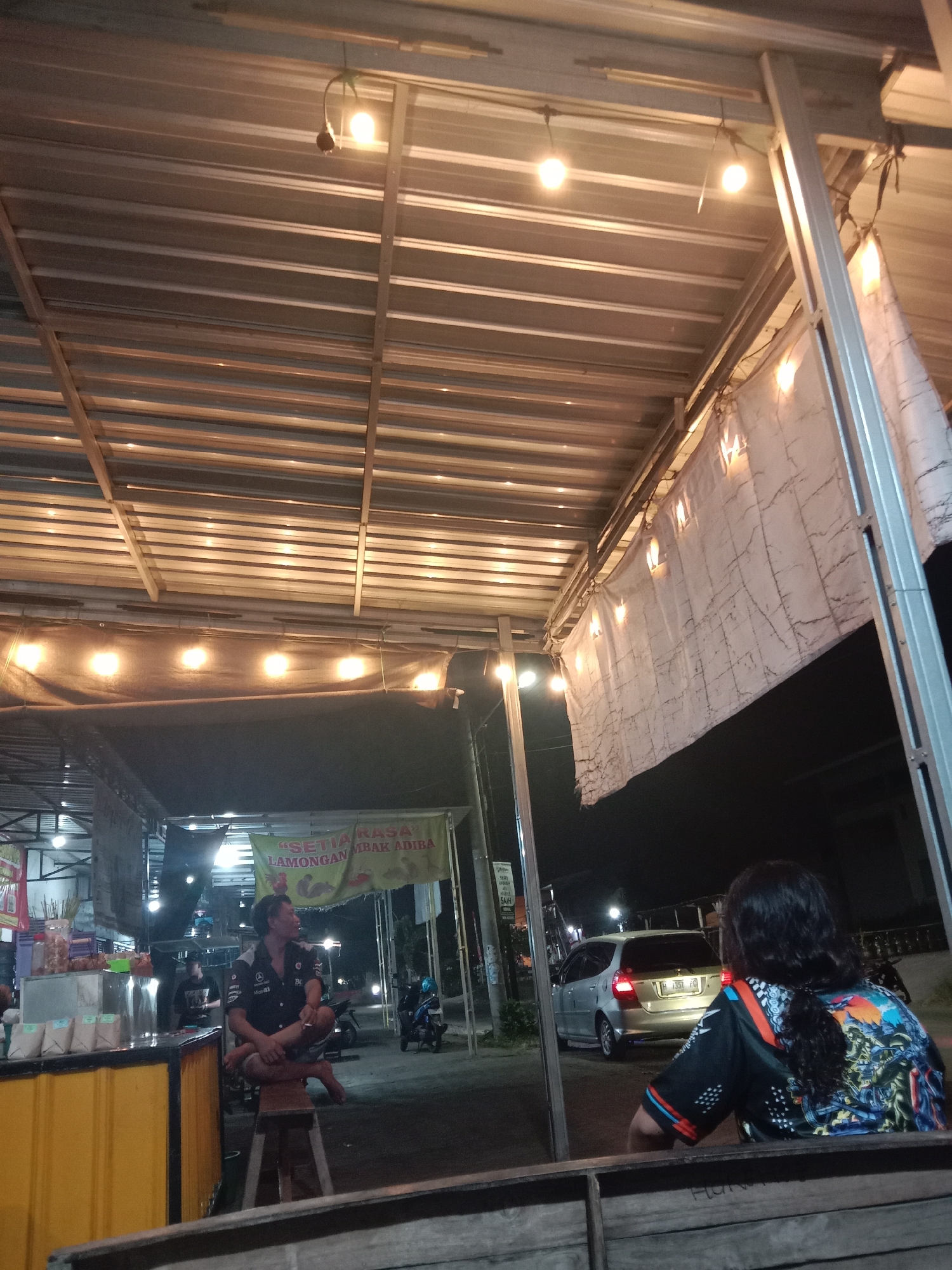 The image depicts an outdoor night scene, likely at a street food stall or small restaurant, with overhead corrugated metal roofing supported by metal beams. String lights illuminate the area, casting a warm glow. In the foreground, a person with long dark hair is seated, viewed from the back, wearing a black shirt with a colorful graphic design on the back. They are looking towards a silver hatchback car with a license plate that appears to read "H 1351 PD". Further into the scene, a man in a dark collared shirt is sitting cross-legged on a wooden stool. Behind him, a yellow food stall counter is visible. A banner with text, including "SETIA RASA LAMONGAN MBAK ADIBA", is hanging above. Various bags and items are displayed on the counter. A motorcycle is parked to the right of the man on the stool, and another motorcycle is visible further in the background with a rider. The environment suggests a casual, potentially busy, evening gathering place in Cangkring, Indonesia.