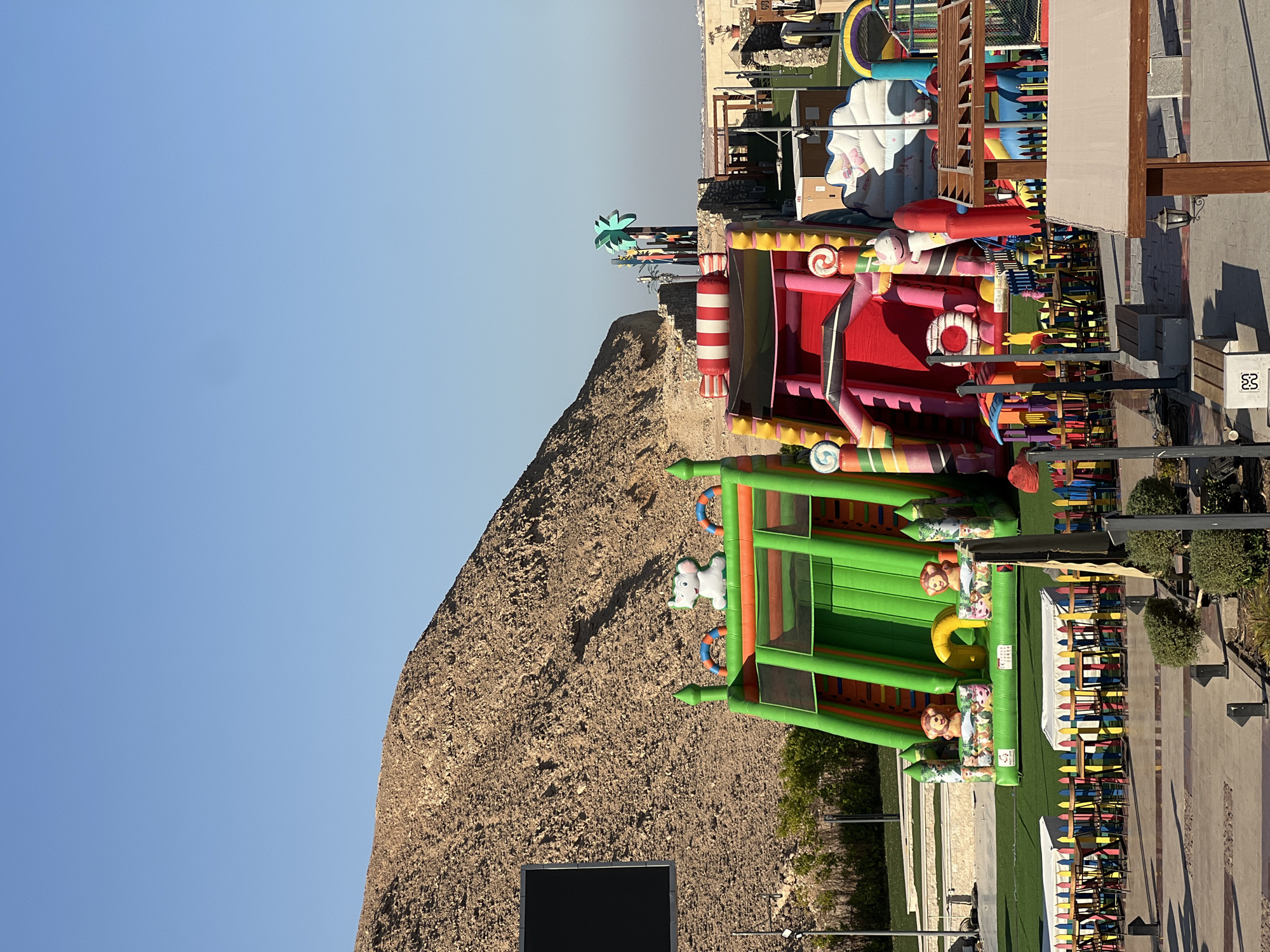 In Sharm El Sheikh, Egypt, a sunlit outdoor recreational area features two large inflatable slides. The slide on the left is predominantly green with orange accents and features cartoon animal graphics at its base, including lions, pigs, and rabbits. A white elephant figure sits atop this slide. The slide on the right is bright pink and red, decorated with candy-like elements such as striped candy canes and swirl lollipops. It also has a white elephant figure.
Behind the inflatables, a substantial, barren, rocky hill forms a dramatic backdrop, characteristic of the desert landscape surrounding the city. To the right, the scene transitions to more structured elements: a fenced-off area with small tables and chairs suggests a seating or dining space. Further back, some palm tree-shaped decorations are visible, hinting at a tropical theme. The overall environment appears to be a clean and well-maintained amusement or park area, likely catering to families. The sky is a clear, bright blue, indicating fair weather.