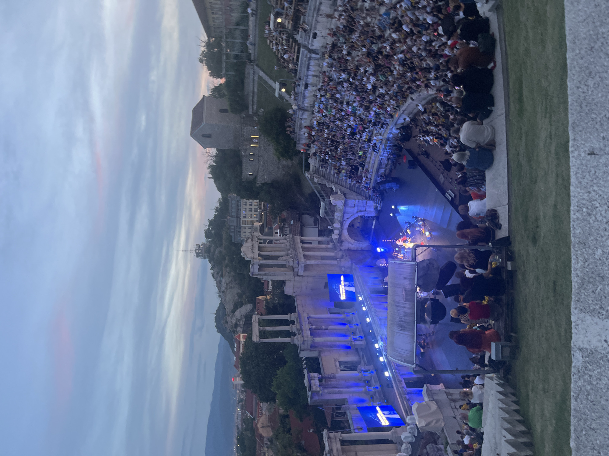 This is a medium-shot, eye-level, outdoor, handheld photograph of an ancient amphitheater at dusk. The amphitheater is filled with a large audience seated on stone tiers. A stage is set up at the bottom of the amphitheater with blue lighting and screens displaying images. Performers are visible on the stage. The sky above is a mix of blue and pink, indicating the time of day is twilight. In the background, there are hills with structures and buildings scattered across them, and mountains in the distance. The foreground shows a grassy area and the edge of a stone seating structure.
