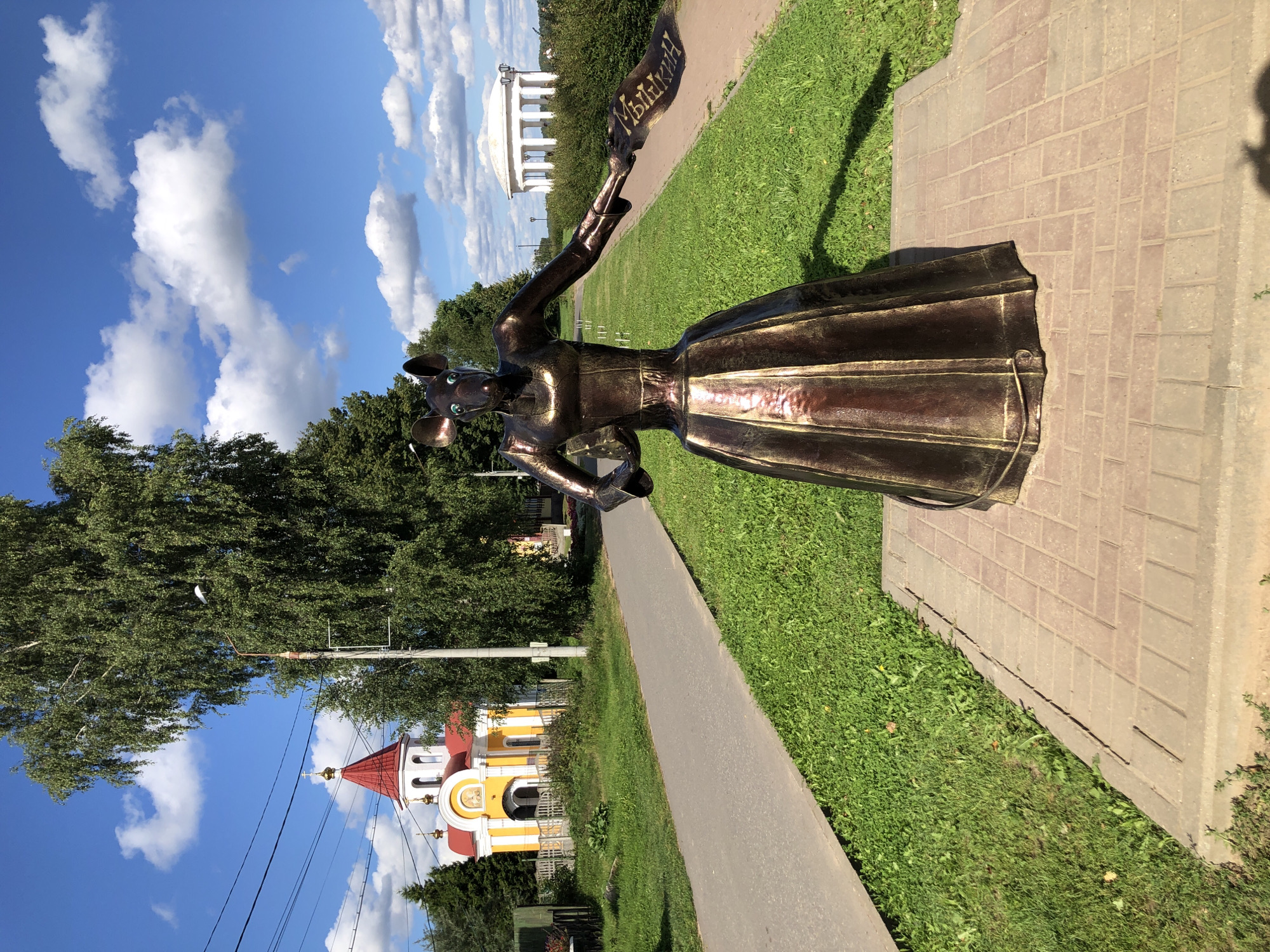 A bronze sculpture of a mouse wearing a dress stands in a paved area next to a grassy lawn. The mouse sculpture holds a banner with Russian text. In the background, a yellow and red church with a domed roof is visible. A white colonnaded structure with a domed roof is also present in the background, along with trees and a blue sky with white clouds. The scene appears to be in the city of Myshkin, Russia, known for its mouse-themed attractions.