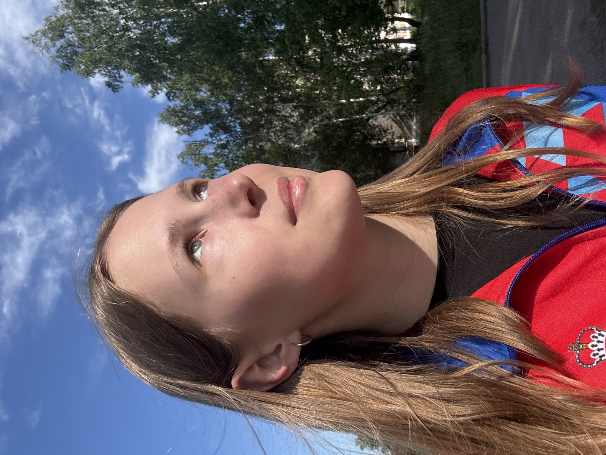 The image is rotated 90 degrees clockwise, depicting a young person looking upwards towards a bright blue sky. The subject is a young individual, likely female, with fair skin, light-colored eyes, an upturned nose, and full, slightly parted lips. Her long, light brown hair frames her face and flows down. A small silver hoop earring is visible in her right ear.
She is wearing a vibrant red jacket with prominent blue and lighter blue checkered or geometric patterns on the sleeves and shoulders, and a black undershirt is visible beneath. On the right side of her chest, an emblem resembling a stylized crown with red, white, and blue elements is clearly visible on the red fabric.
The background features a clear, deep blue sky with a few wispy white clouds. Lush green foliage of trees is visible along the upper edge of the image (when oriented correctly), suggesting an outdoor setting like a park or natural area. The bright sunlight creates strong highlights on her hair and skin, indicating a sunny day. The scene evokes a calm, contemplative mood under an open sky in Chaikovsky, Russia.