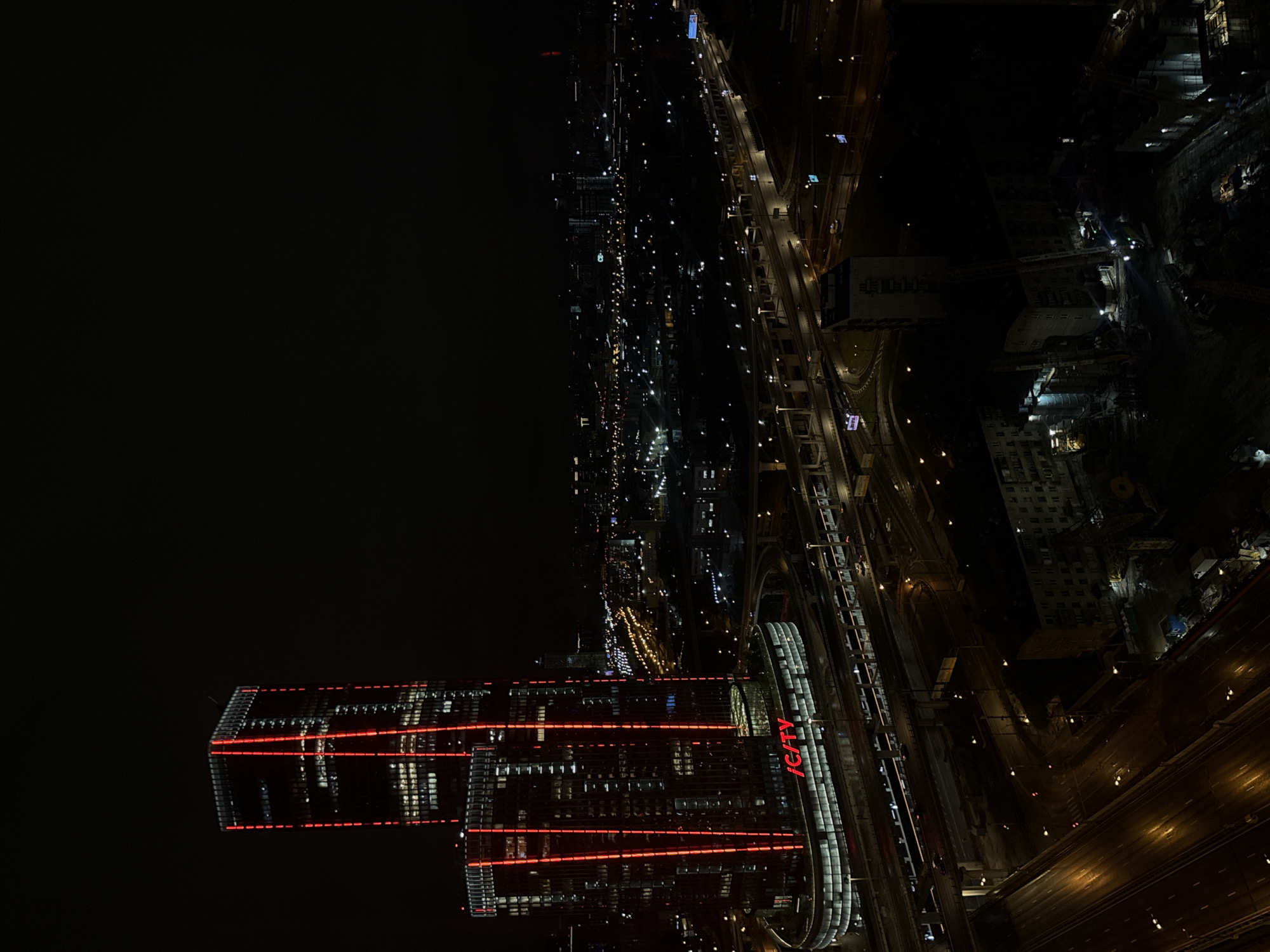 The image depicts an aerial view of a cityscape at night, likely in Khimki, Russia. The dominant feature is a complex of modern buildings, illuminated with red accent lights, with the "ICITY" logo prominently displayed. These buildings are located adjacent to a railway line and highway, both brightly lit. Further into the background, the city stretches with sparse yet pervasive lights, indicating residential and commercial areas. The sky is dark and mostly devoid of detail.