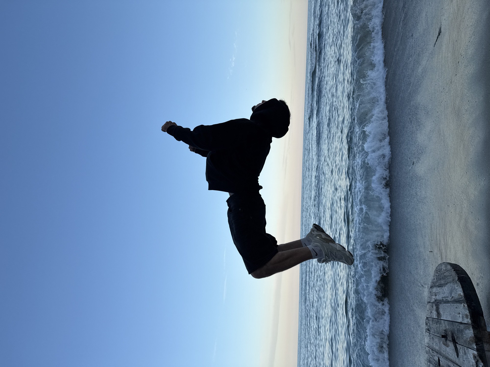A person in a black hoodie and shorts is captured mid-jump against a backdrop of a beach scene. The location appears to be on a beach in Varna, Bulgaria, evidenced by the presence of the Black Sea, sandy shore, and a wooden cable spool typical for coastal areas. The sky transitions from a clear blue to a lighter hue near the horizon, indicating a sunrise or sunset. The individual is silhouetted, their face obscured by the hood. Their arms are raised, and they are wearing light-colored sneakers. Waves are visible near the shoreline. The image is rotated 90 degrees counterclockwise.