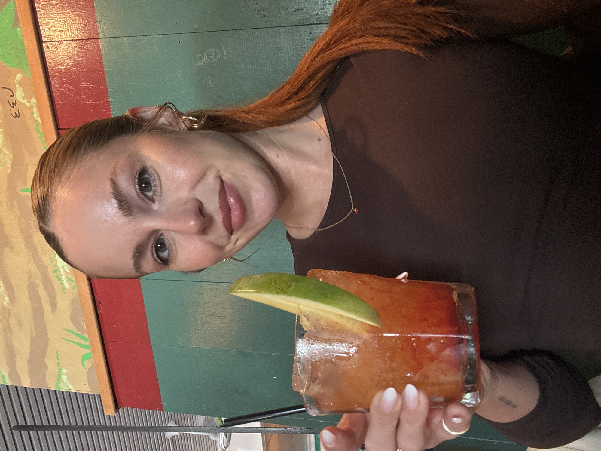A woman with red hair, wearing a brown top and a delicate gold necklace, is holding a cocktail in a glass with a lime wedge. The cocktail has a reddish-orange hue. The woman is smiling and looking at the camera. Her fingernails have an ombre effect with white tips. The background features a green and red painted wooden wall. The location is likely in Los Angeles, United States of America.