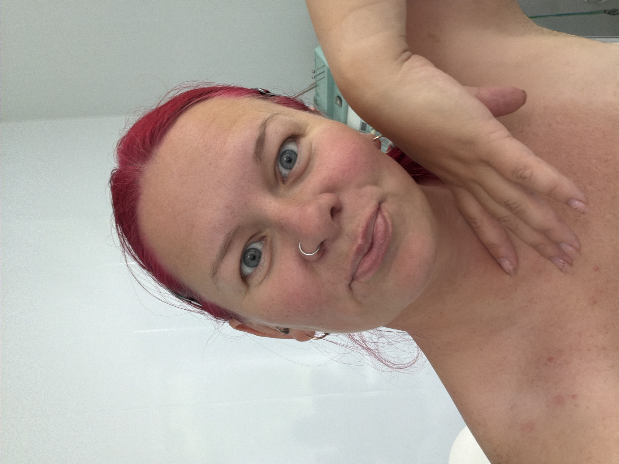 The media file features a close-up selfie of a woman with distinct bright pink/red hair and piercing blue eyes. She has a silver hoop nose ring and small hoop earrings. Her head is tilted, and she looks directly at the camera with a subtle, playful smirk. Her fair skin shows visible freckles and some minor blemishes. One of her arms is raised, and her hand, with lightly polished nails, rests gently on her shoulder, which is partially exposed.
The setting appears to be an indoor room, likely a bathroom, characterized by plain white walls or tiles in the background, suggesting a clean and minimalist environment. Faint reflections of what might be a light fixture or a cabinet are discernible in the blurred background. The overall lighting is bright and even, consistent with indoor illumination. The activity captured is the woman posing for a personal photograph. The location context for this scene is Chaikovsky, Russia. There is no visible text in the image.