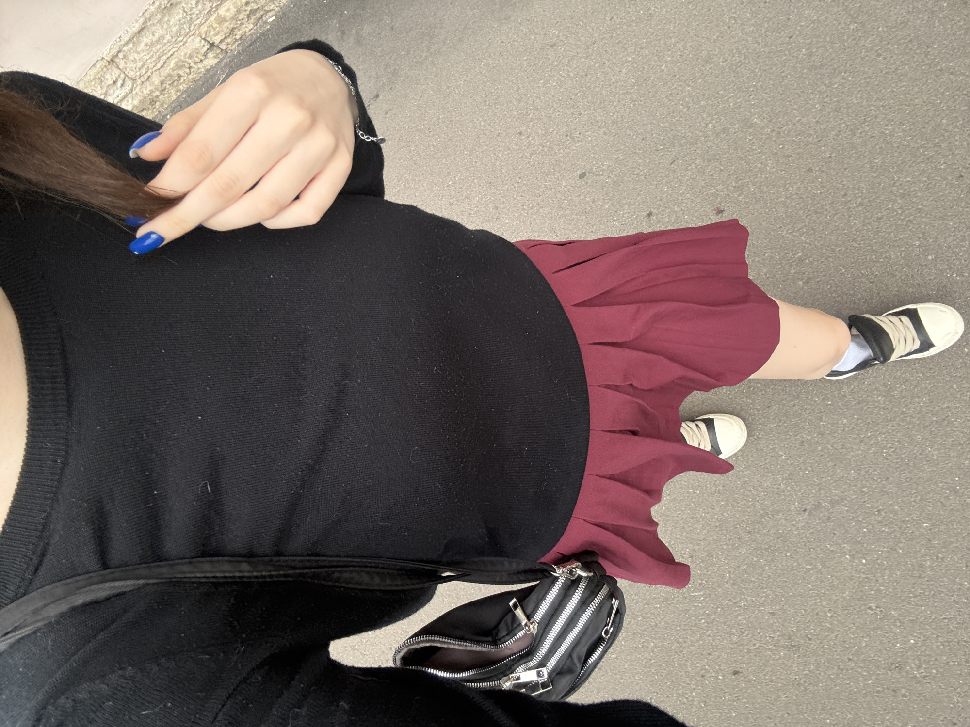 A first-person perspective shot from above shows a person standing on asphalt outdoors, possibly in Saint Petersburg, Russia. The person is wearing a black long-sleeved shirt, a maroon pleated skirt, and black and white sneakers with white socks. A black crossbody bag with multiple zippered compartments is slung over the person's shoulder. The person's right hand is raised, and their fingers are adorned with bright blue nail polish. The background consists of a textured concrete wall and the asphalt ground.