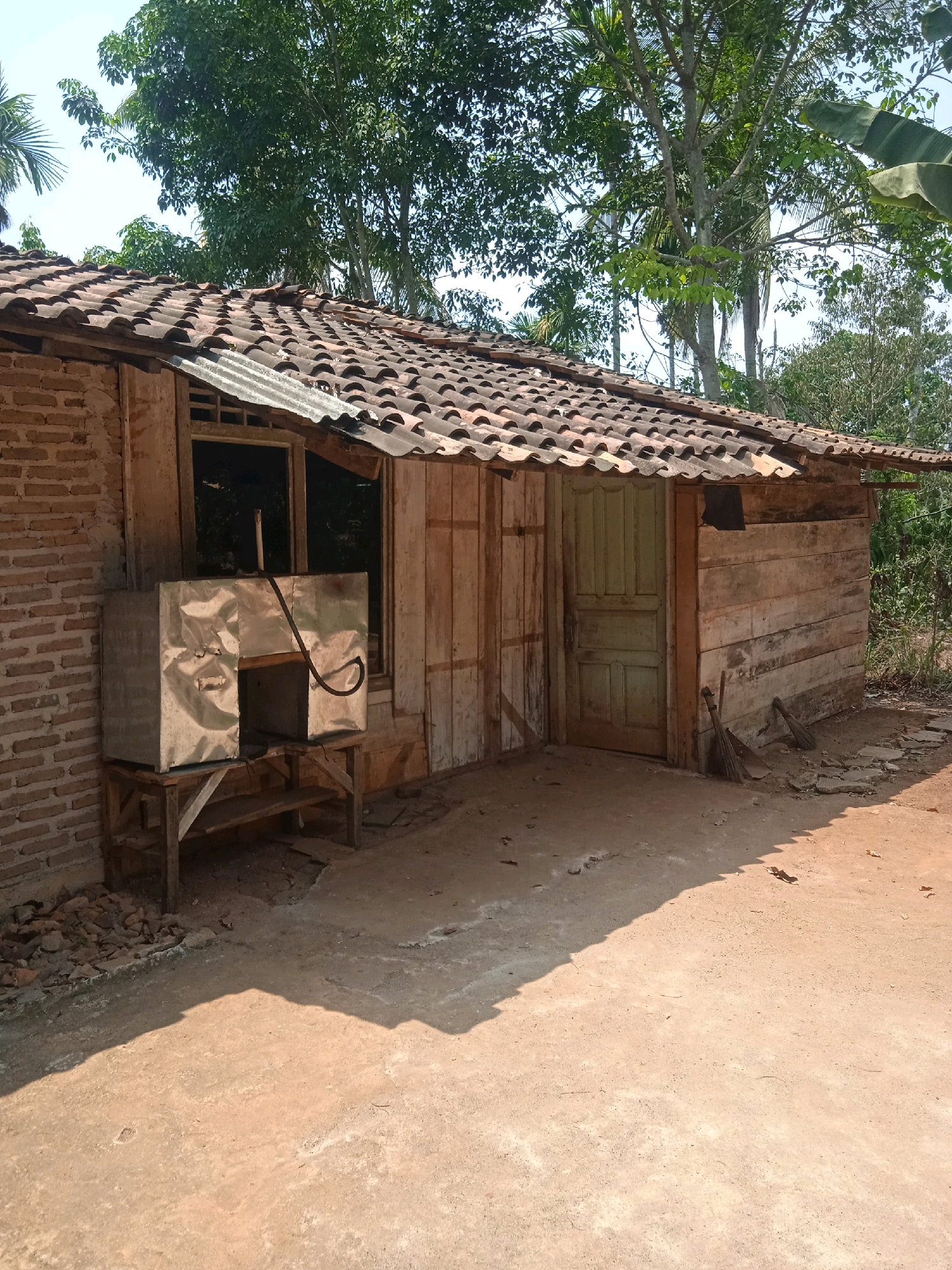 The image depicts a rustic, single-story structure with a corrugated tile roof and wooden plank walls. A brick section forms one side of the building, featuring a window with a metal enclosure. In front of the window sits a stainless steel contraption on a wooden stand, with a dark hose extending from it. Adjacent to the main structure is a green wooden door. Two brooms lean against the right side of the building. The ground in front is a dirt and concrete surface, with shadows cast by the structure and trees. Lush green trees and vegetation surround the building, suggesting a rural or semi-rural environment. The scene appears to be an outdoor area in Karang Sari, Indonesia.