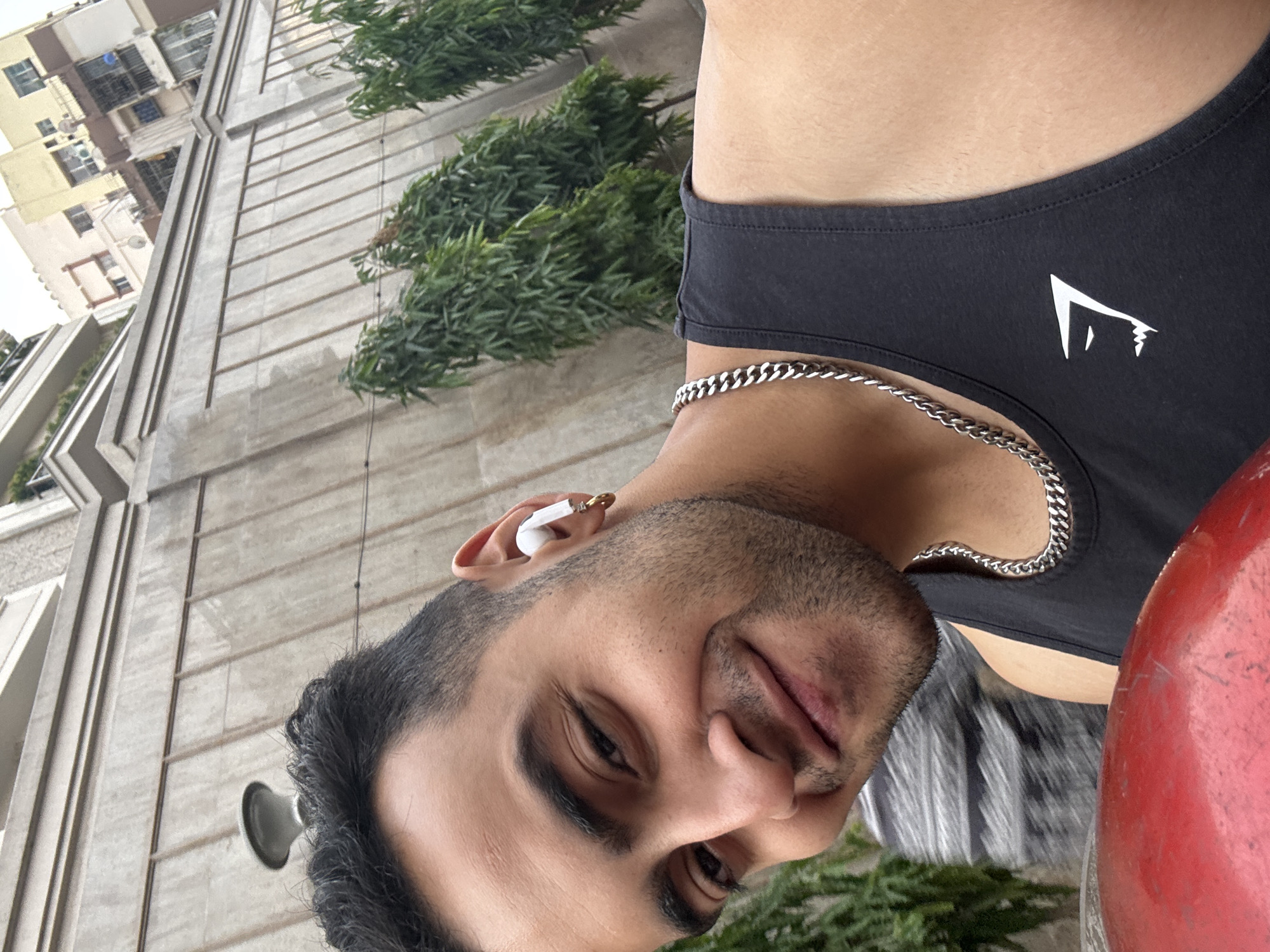 A selfie image of a man in Kolkata, India. The man is wearing a black tank top with a white logo on the chest, and a silver chain around his neck. He has short black hair and is wearing a white wireless earbud in his left ear, with a gold hoop earring. He is smiling slightly. Behind him, there is a concrete building with green foliage and a red bollard. Other buildings are visible in the background.