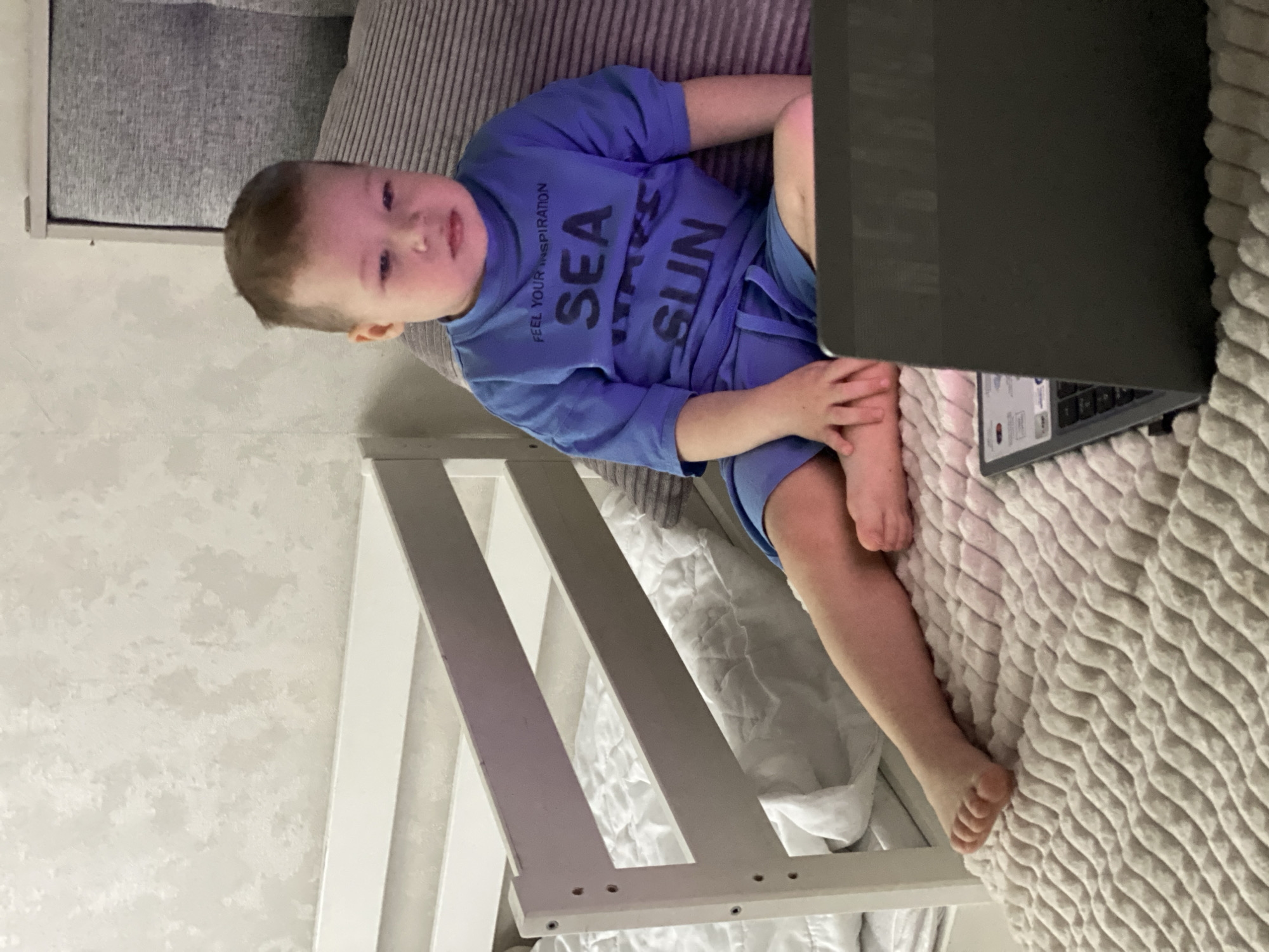 A young child sits on a patterned bed, resting against a gray striped pillow. The child is wearing a blue T-shirt with "SEA SUN" printed on it and matching blue shorts. A partially visible laptop is positioned in front of the child. A white cot or crib frame is visible behind and to the left of the child. The location cannot be determined from the image.