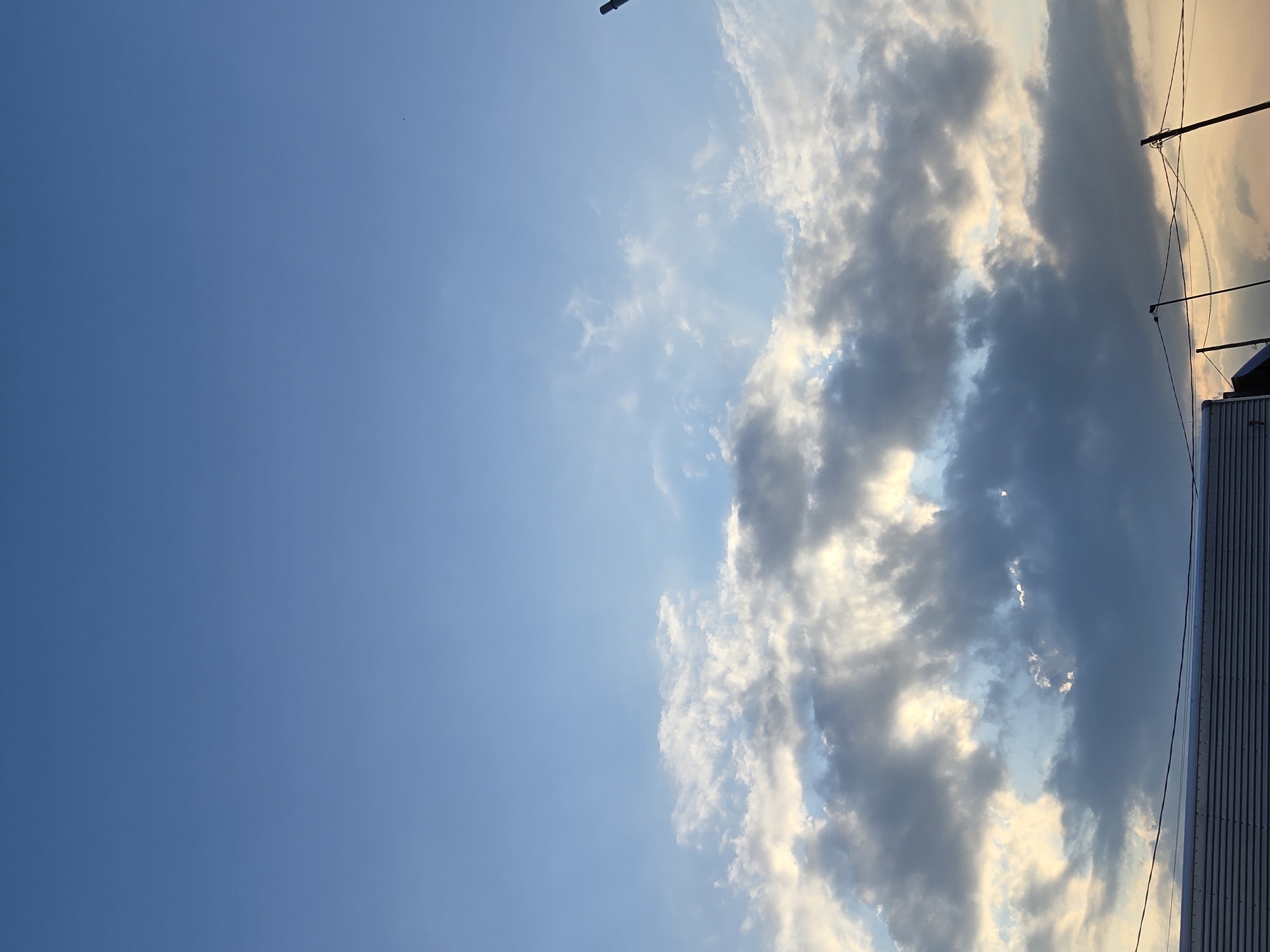 The image captures a twilight sky over a low-rise urban landscape, likely in Ермолаево, Russia. The sky is a gradient of clear, pale blue at the top, transitioning to lighter hues with wispy, illuminated clouds in the middle. Dense, darker clouds are prominent in the lower half of the sky, with shafts of sunlight breaking through them. In the foreground, the roofline of a building is visible, characterized by horizontal ribbed panels, suggesting a modern construction. Power lines are strung across the scene, supported by poles, and a dark, cylindrical object, possibly a light fixture or utility pole, is visible on the right. The overall impression is of a calm, late evening in an inhabited area.