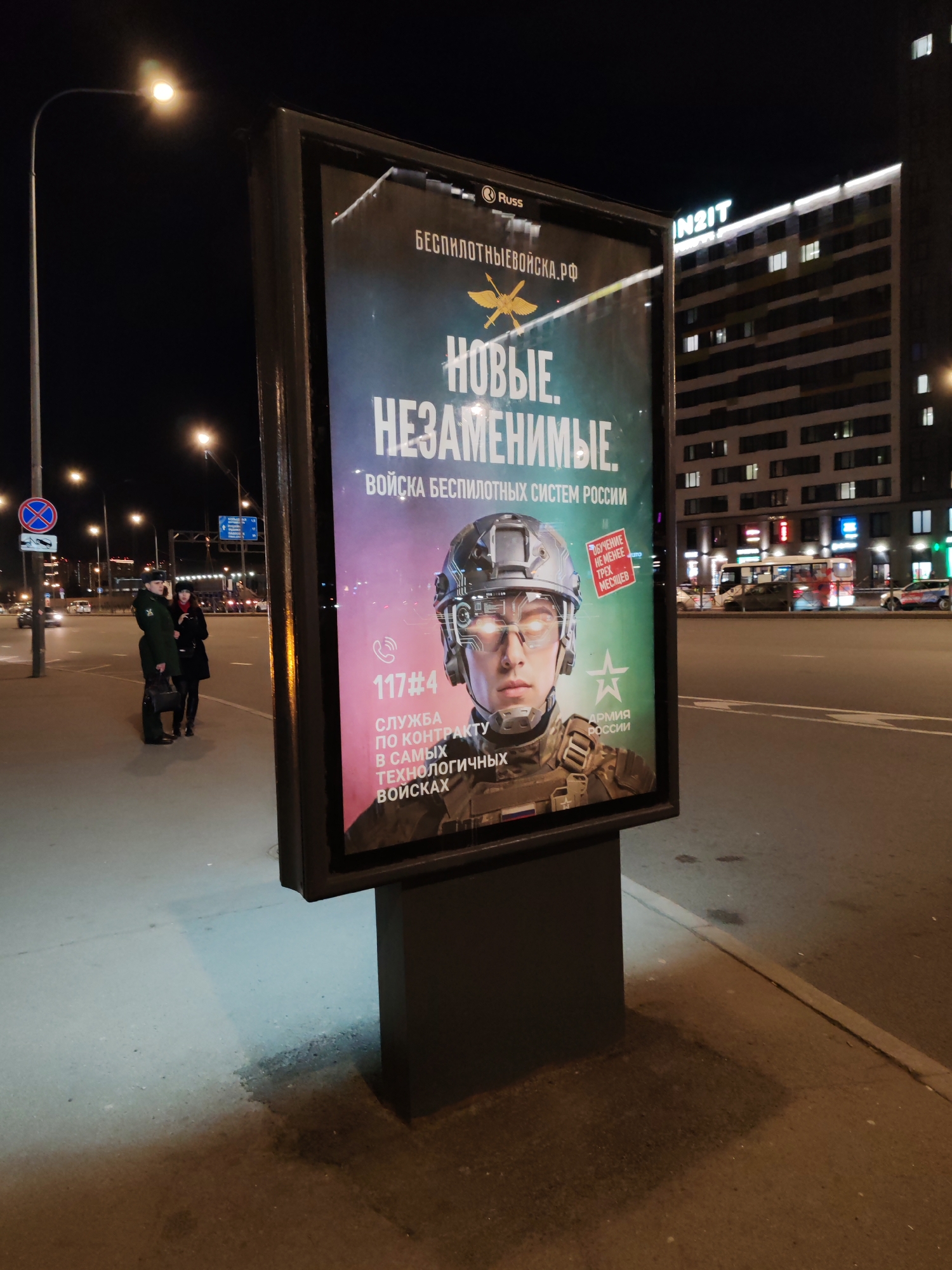 The image depicts a nighttime urban scene in Saint Petersburg, Russia, centered around a prominent illuminated billboard on a street sidewalk.
The billboard, branded with "Russ," is an advertisement for military recruitment. Its main text reads "НОВЫЕ. НЕЗАМЕНИМЫЕ." (NEW. IRREPLACEABLE.) and "ВОЙСКА БЕСПИЛОТНЫХ СИСТЕМ РОССИИ" (Troops of Unmanned Systems of Russia). A website, "БЕСПИЛОТНЫЕВОЙСКА.РФ" (DRONEFORCES.RF), is displayed at the top. The visual features a digitally rendered image of a young man, presented as a soldier, wearing a futuristic helmet and glowing eyeglasses, emphasizing advanced technology. Further text states "ОБУЧЕНИЕ НЕ МЕНЕЕ ТРЕХ МЕСЯЦЕВ" (TRAINING NO LESS THAN THREE MONTHS) and invites for "СЛУЖБА ПО КОНТРАКТУ В САМЫХ ТЕХНОЛОГИЧНЫХ ВОЙСКАХ" (CONTRACT SERVICE IN THE MOST TECHNOLOGICAL FORCES). A contact number "117#4" is provided, along with the "АРМИЯ РОССИИ" (ARMY OF RUSSIA) logo.
In the background, a street with vehicles and streetlights is visible. To the left of the billboard, two individuals stand on the sidewalk; one appears to be in military uniform holding a bag, alongside another person in civilian attire. A "No Stopping" traffic sign is also present on a pole. Across the street, a multi-story building with illuminated windows displays the text "IN2IT." The overall setting is a clear night in a bustling city.