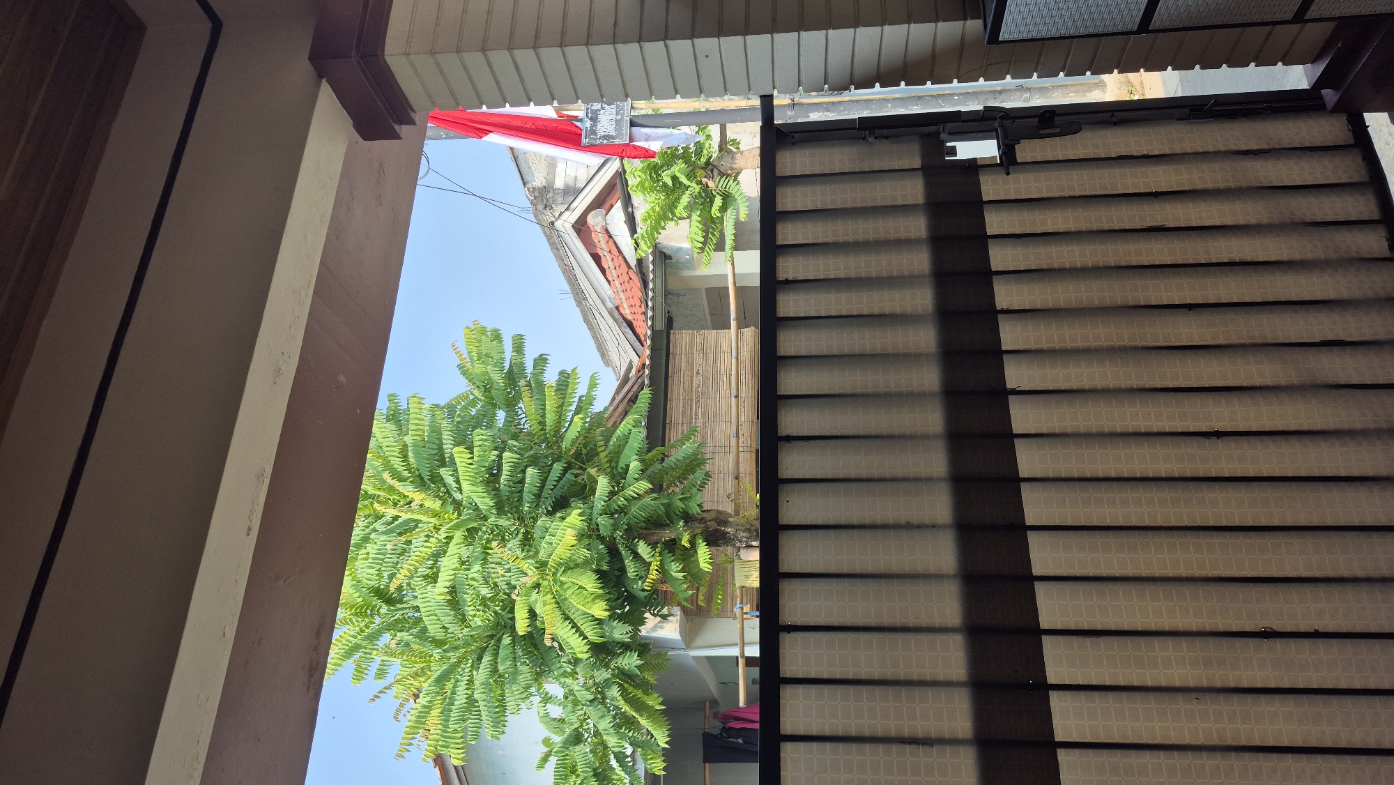 The image shows a view through a patterned metal gate towards a residential area. In the foreground, the gate has vertical slats with a repeating checkered pattern. A shadow from an overhead structure casts diagonal lines across the gate. To the right of the gate, a section of a building column and a white wall are visible.
Beyond the gate, a lush green tree with compound leaves dominates the center of the frame. Behind the tree, there are houses with pitched roofs, one with red tiles. A bamboo-like screen is visible on one of the buildings. To the far right, an Indonesian flag, red and white, is affixed to a pole attached to a building column. The sky is clear and blue. The scene appears to be in a residential neighborhood in South Tangerang, Indonesia, likely during daylight. There are no people visible in the image.