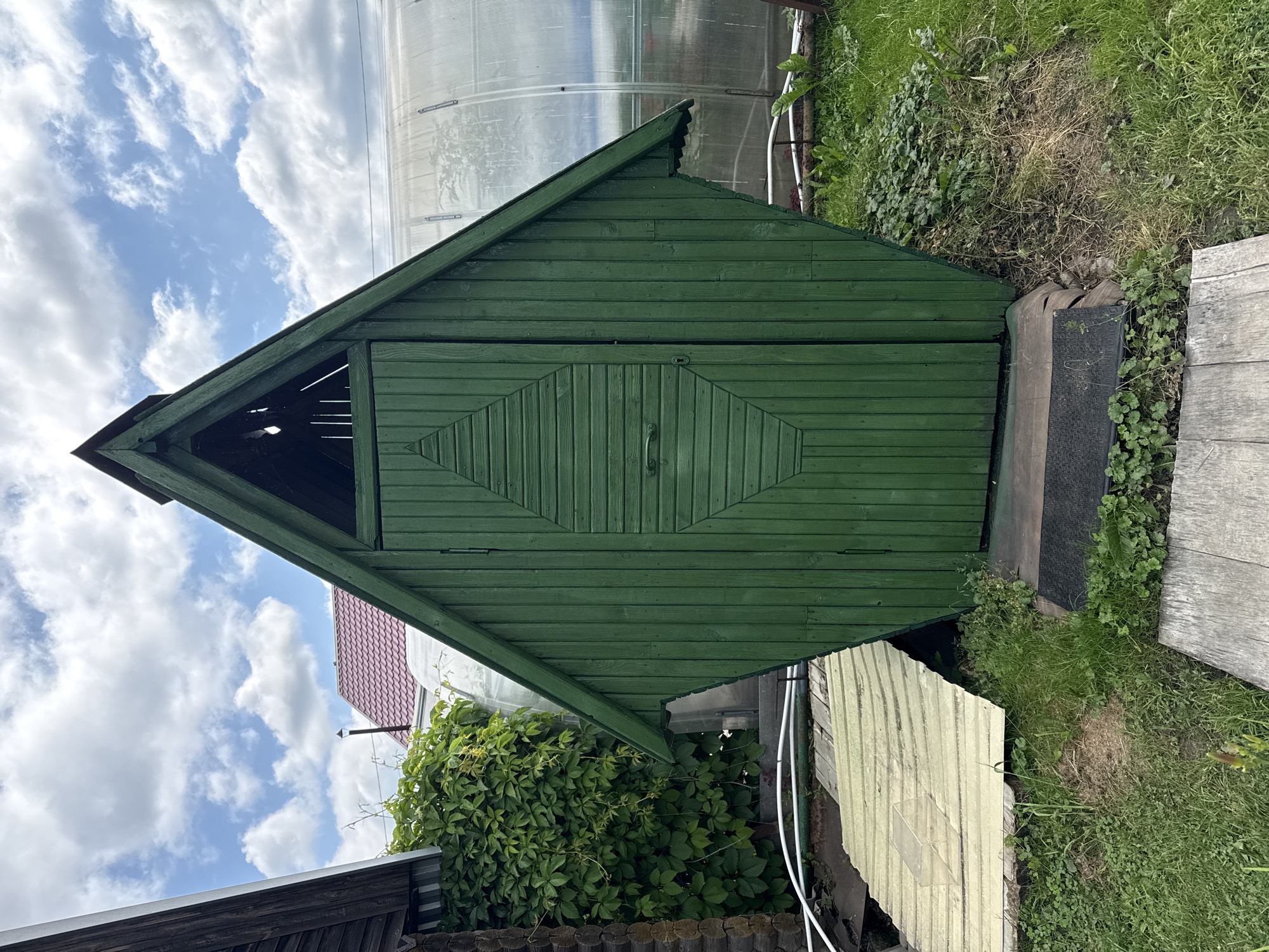 The image depicts an outdoor scene in Baryshevo, Russia, featuring a small, dark green wooden shed with a distinctive diamond-shaped panel on its door. The shed has a pointed roof. To the left of the shed, a section of a wooden building and a dense wall of green ivy are visible. A transparent, arched greenhouse structure is situated to the right of the shed. The ground around the shed is covered in grass and some debris. A light-colored corrugated panel rests on the ground in front of the shed, and a wooden path leads towards it. The sky above is partly cloudy with white clouds against a blue backdrop.