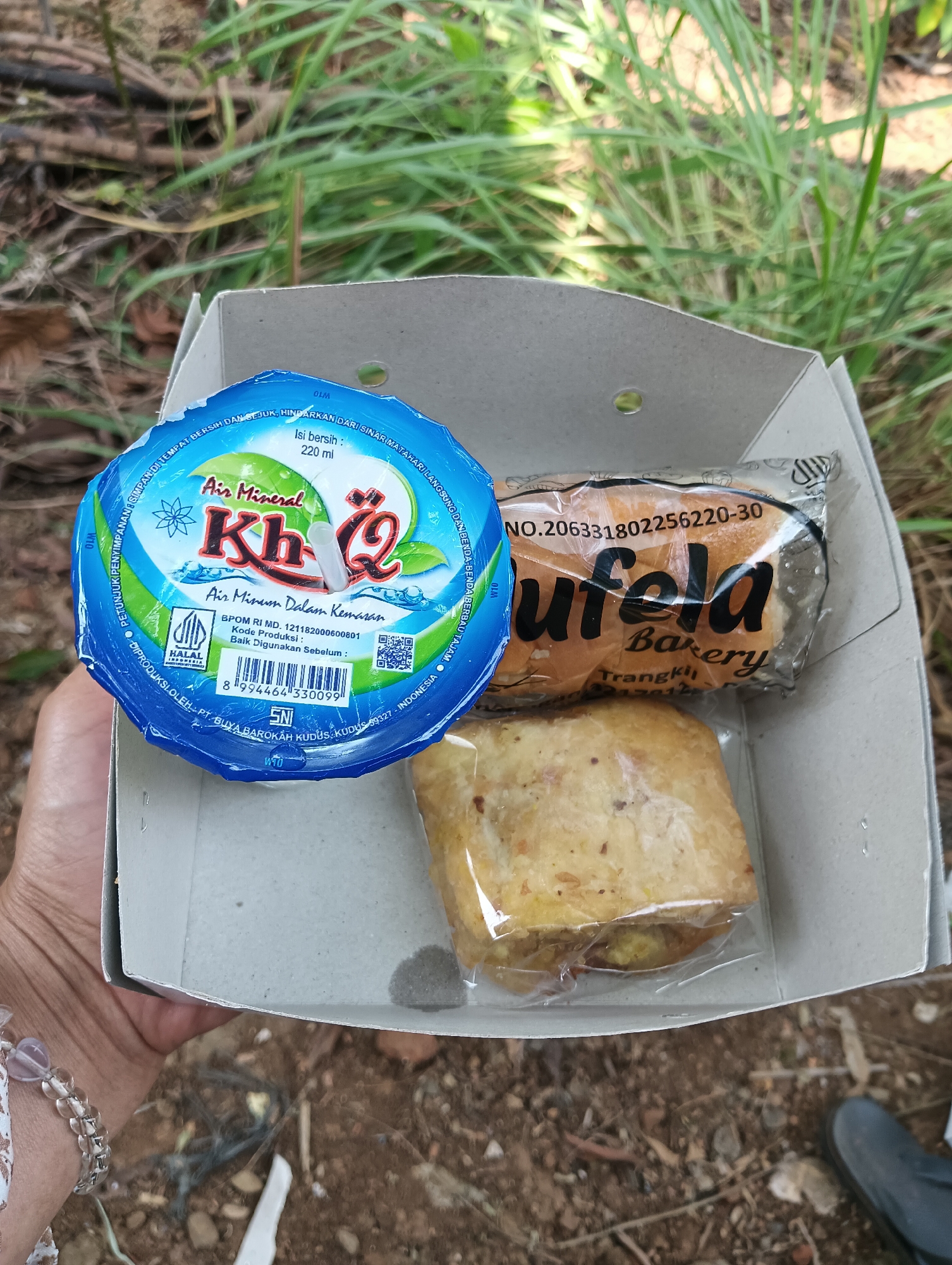 A person's hand, adorned with a bracelet, holds an open cardboard food container filled with snacks. The container contains a blue-capped bottle of "Air Mineral Kh-Q" with a white straw, a plastic-wrapped pastry labeled "Nufela Bakery," and another pastry wrapped in clear plastic. The background shows dry grass and soil, suggesting an outdoor setting, possibly near Cluwak, Indonesia, given the local branding on the products. The lighting indicates it is daytime.