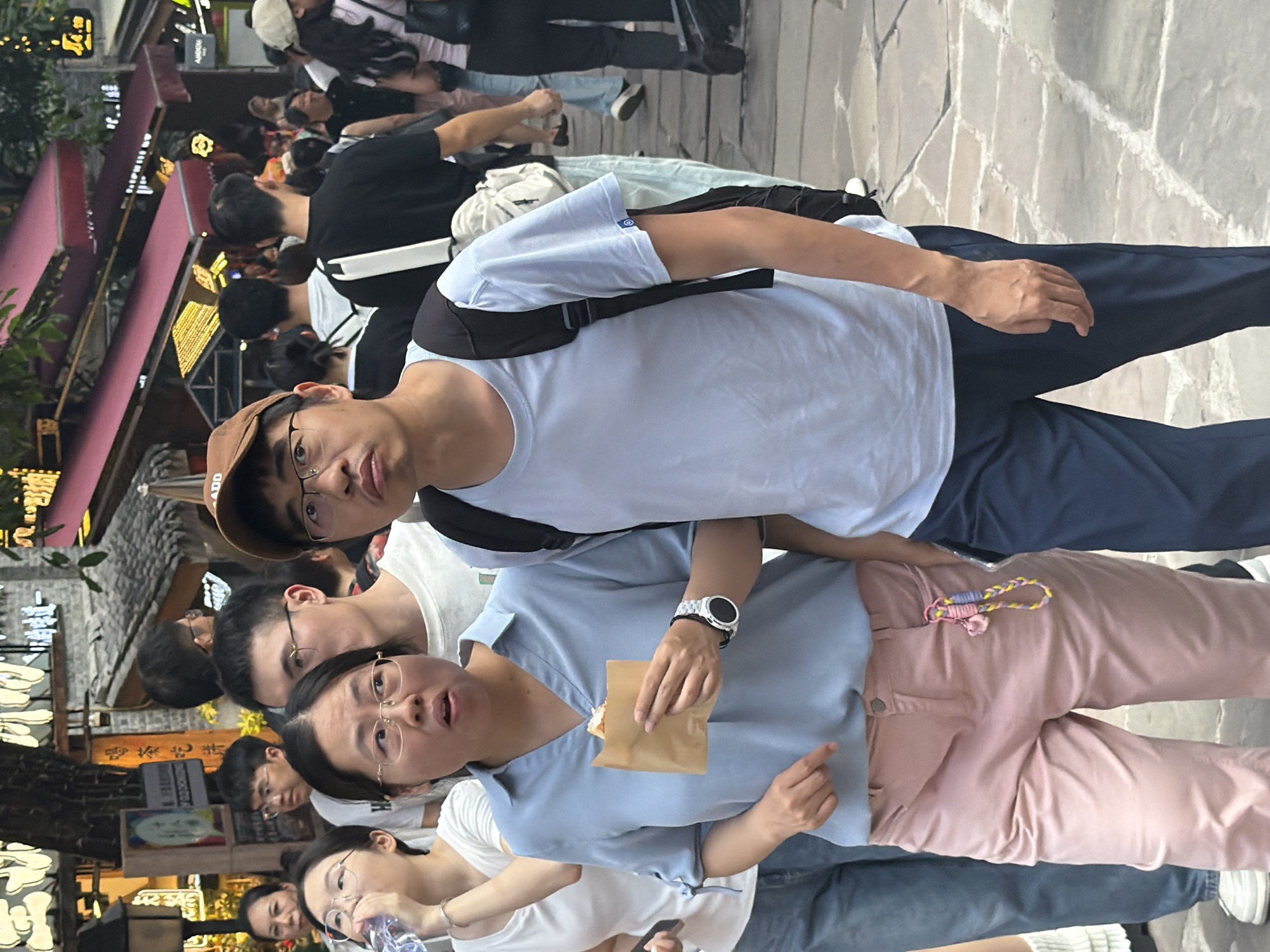 The image shows a group of young people in what appears to be a busy outdoor market or street. The perspective is a close-up, taken from a low angle, looking up at the subjects. One person in the foreground, wearing a light blue t-shirt and a brown cap, is looking directly at the camera. To their right, another person in a light blue t-shirt is holding a small paper package and looking slightly to the side. A third person, wearing glasses and a white t-shirt, is also visible in the foreground.
In the background, more people are seen walking through the street. They are dressed in casual clothing. The setting appears to be a commercial area with awnings, signs, and cobblestone pavement. Some of the signs have Chinese characters, indicating the location is likely in China, consistent with the Yuzhong District context. The lighting suggests it is daytime, possibly late morning or afternoon. The atmosphere seems lively and casual, with people going about their day. The people in the foreground appear relaxed and engaged, with some looking at the camera. There are no obvious signs of a specific event, but rather a snapshot of everyday life in a bustling urban environment.