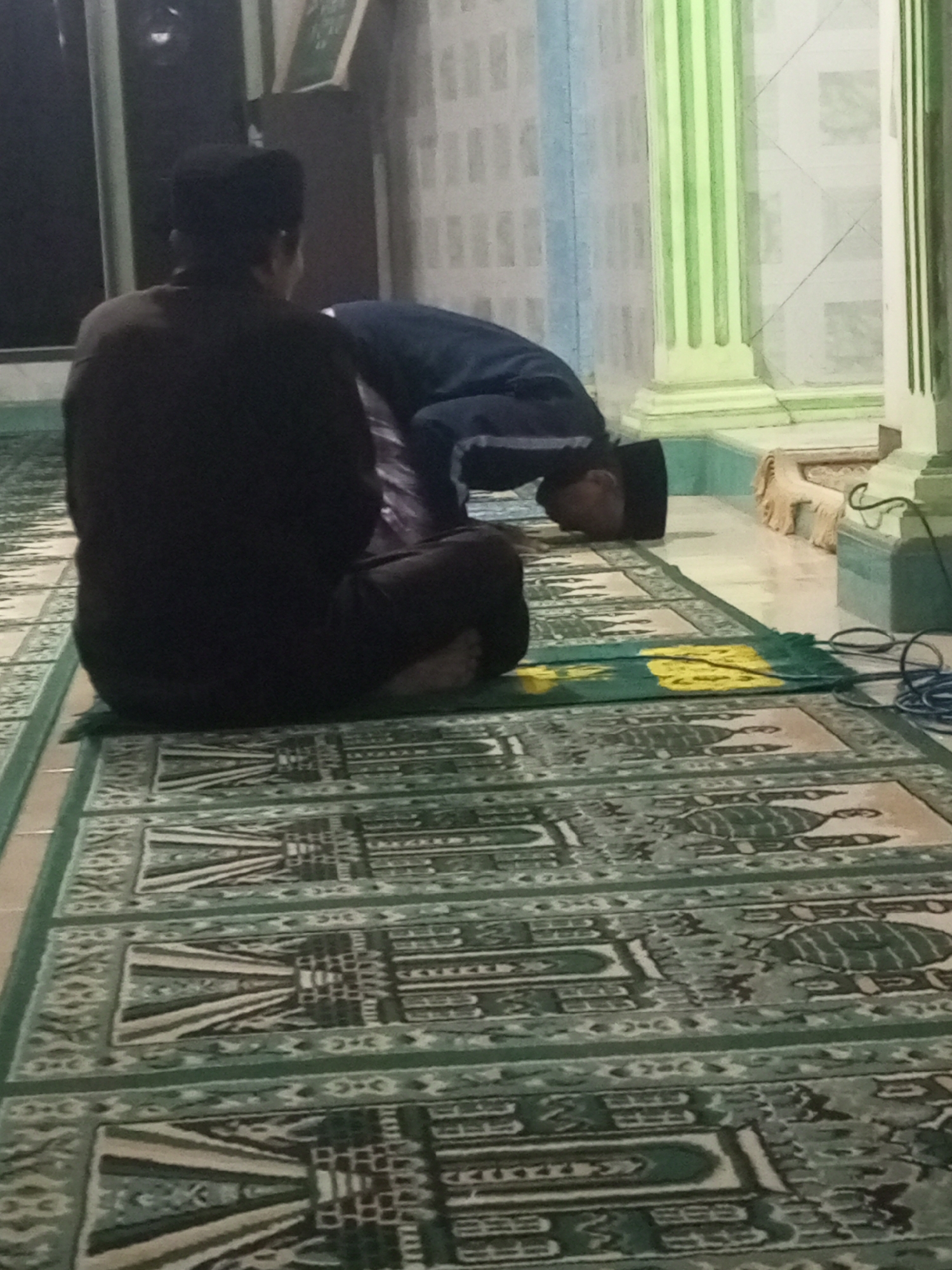 The image depicts two men engaged in prayer on prayer mats inside a building, likely a mosque, in Andongsili, Indonesia. The man in the foreground, viewed from behind, is seated facing away from the camera. He is wearing a dark brown shirt and dark pants, and a black cap. To his right, a second man is in a prostrated position, with his forehead touching a prayer mat. He is wearing a dark blue long-sleeved shirt and dark pants, also with a black cap. Both men are positioned on elaborately patterned green prayer rugs laid out on a tiled floor. The background reveals architectural elements, including columns painted in a light green hue and patterned walls. A green and yellow prayer mat is also visible between the two men. The lighting suggests an indoor setting, possibly during evening prayer.