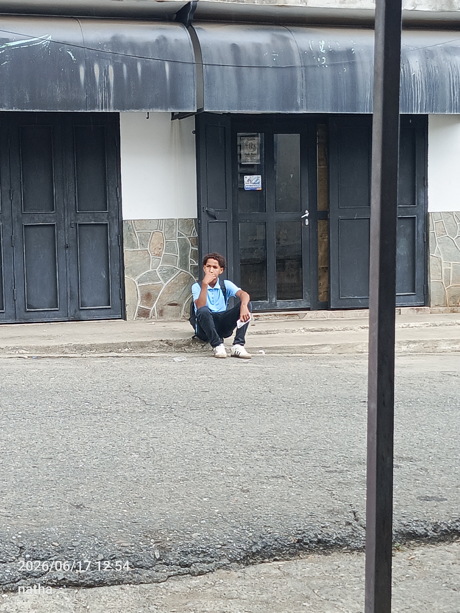 A young man wearing a light blue polo shirt and dark pants sits on a concrete curb outside a building in San Sebastián, Venezuela. He has a dark backpack on his back and is looking to his right with his chin resting on his hand. The building behind him has large, dark double doors and a grey awning. The street in front of him is asphalt with some cracks and gravel. The timestamp "2026/06/17 12:54" and the text "natha" are visible in the bottom left corner of the image.
