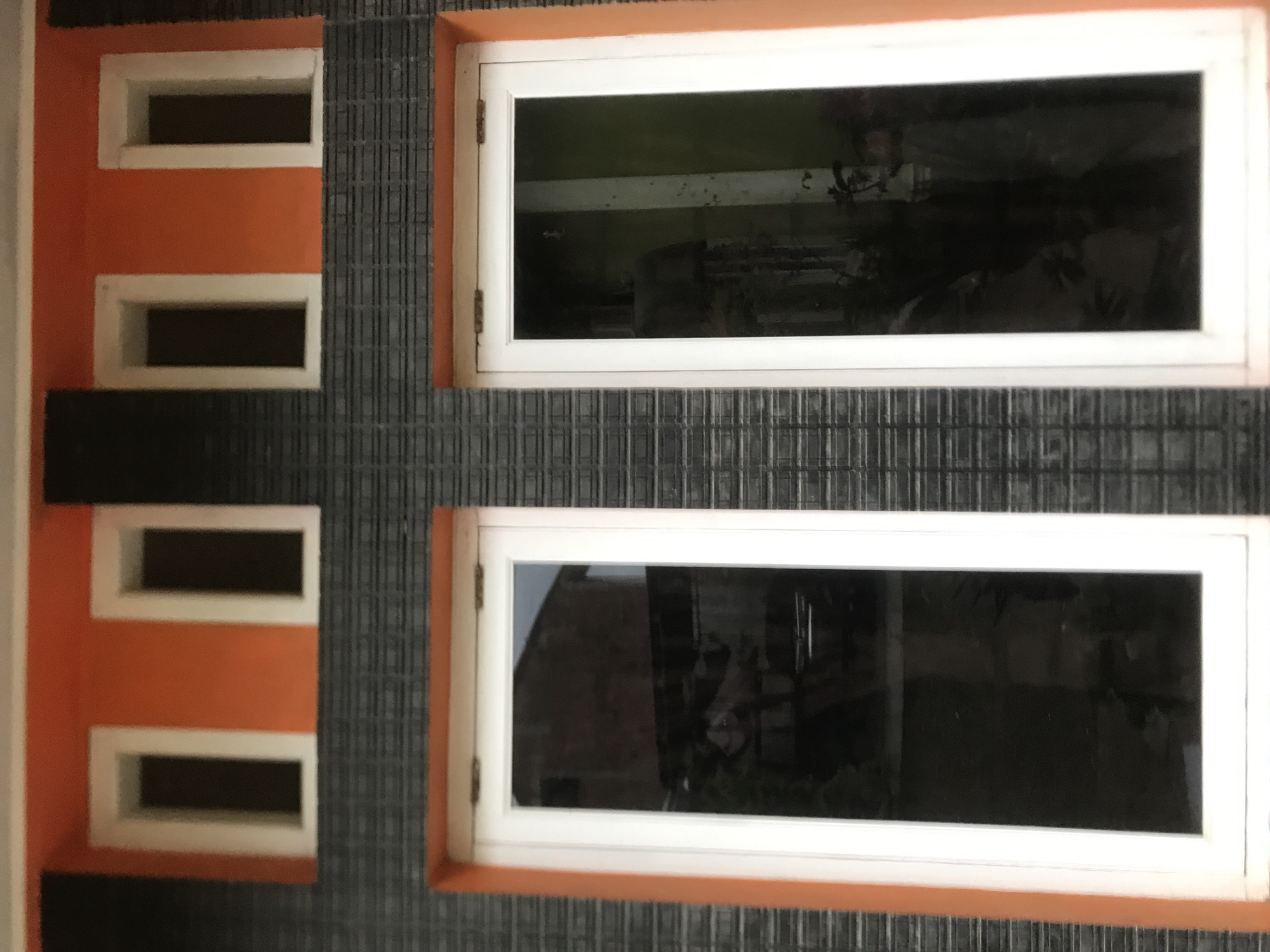 The image depicts a portion of a building exterior. On the left is a section with an orange facade and four small, rectangular window openings. Adjacent to this section is a wall covered in dark gray, horizontally-oriented tiles. To the right of this tiled area are two large, rectangular windows with white frames and orange trim. The windows reflect distorted images of the surrounding environment, obscuring details of what lies inside. The image lacks specific identifying features to pinpoint the precise location within Lhokseumawe, Indonesia.