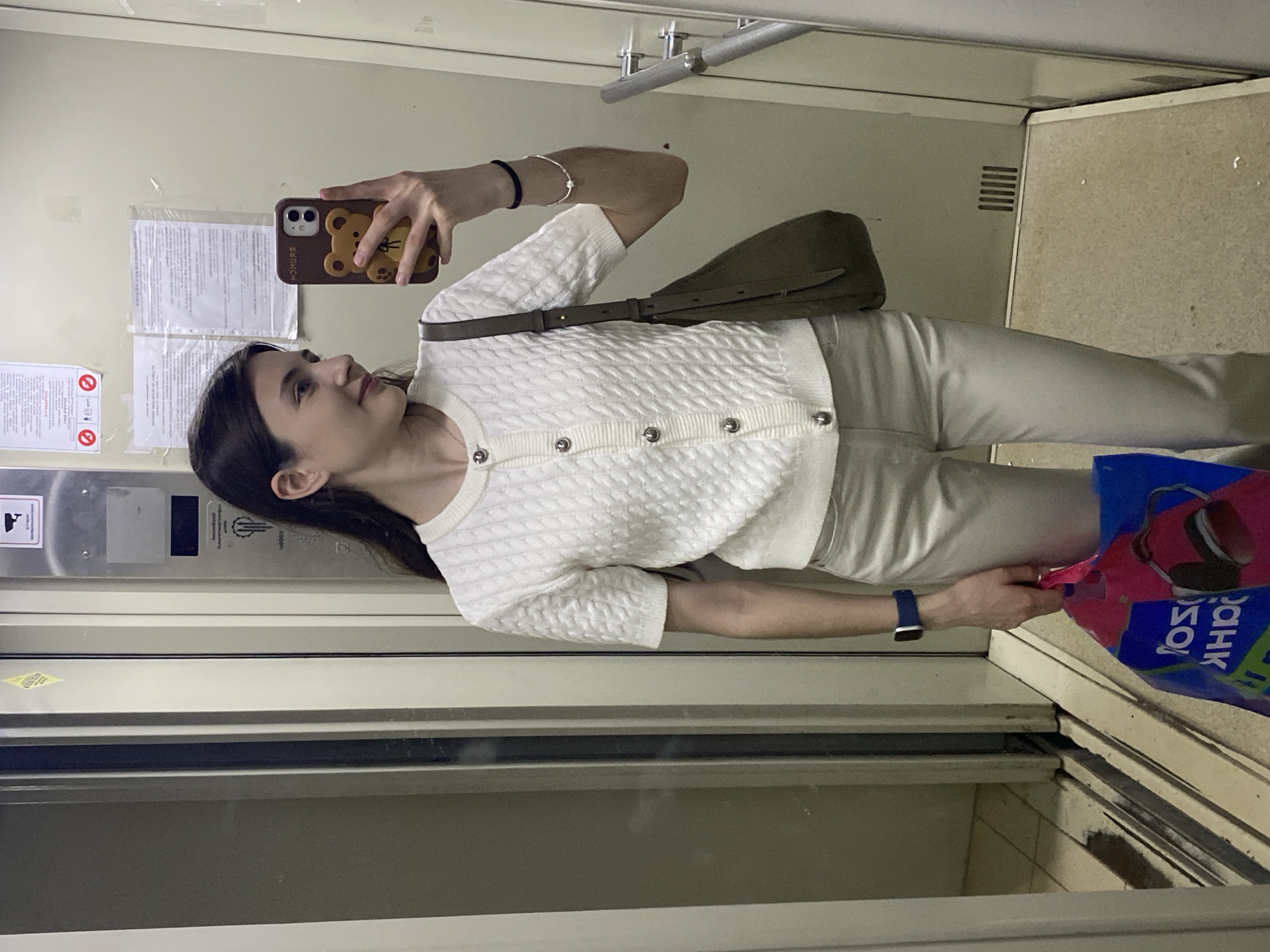 A person is taking a selfie in an elevator. The person has dark hair and is wearing a white cardigan, beige pants, and an Apple Watch. They are holding a brown phone with a bear-shaped case. A green bag is slung over their shoulder, and they are holding a blue shopping bag with red and white text and images. The elevator has metal doors, a control panel, and informational notices. The scene is inside an elevator, which is a common feature in buildings in Votkinsk, Russia, and other cities. It's not possible to determine the location in Votkinsk or the specific type of building from this image.